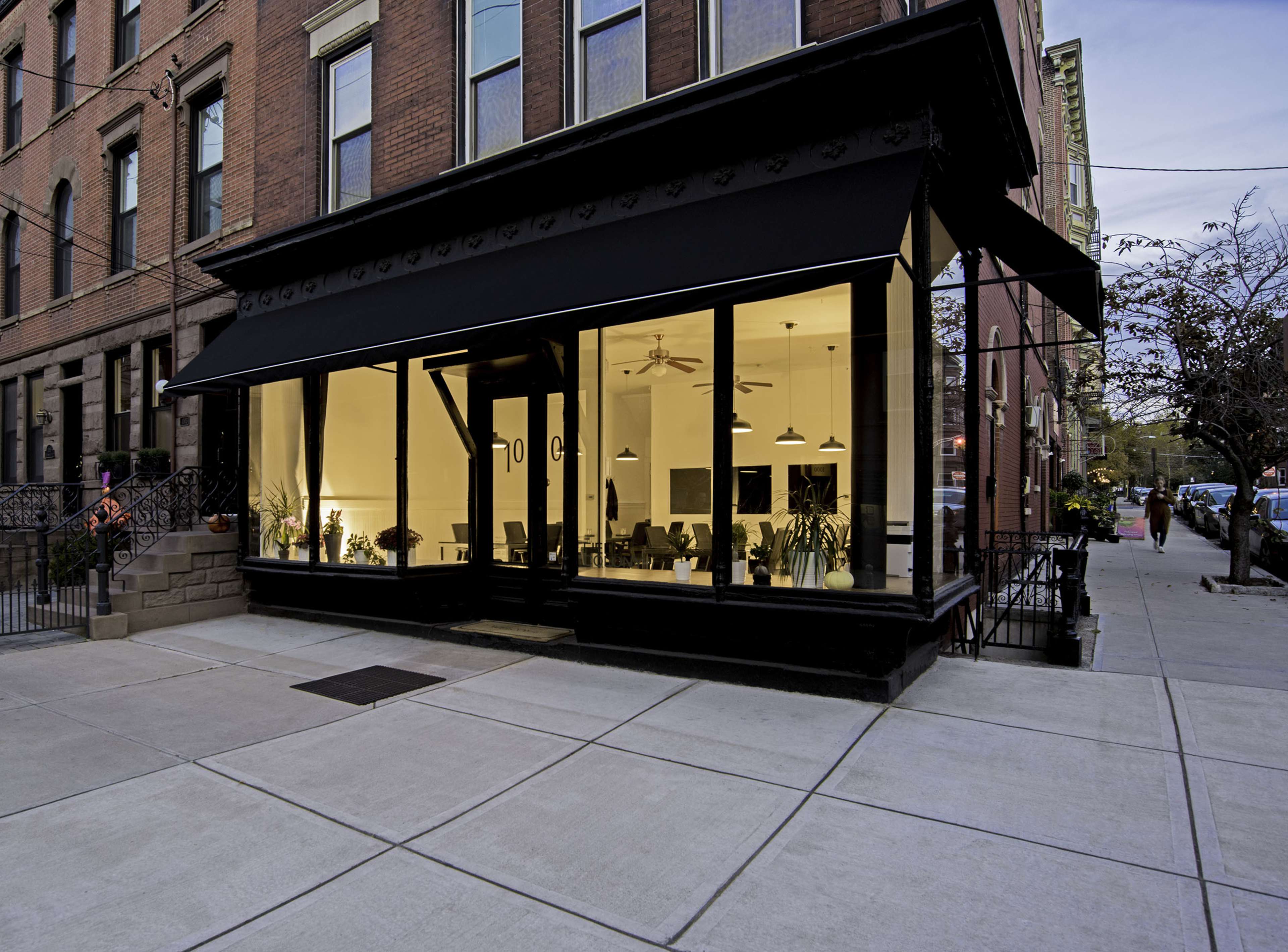 A modern storefront with large glass windows displaying an interior filled with plants, located on a city sidewalk.