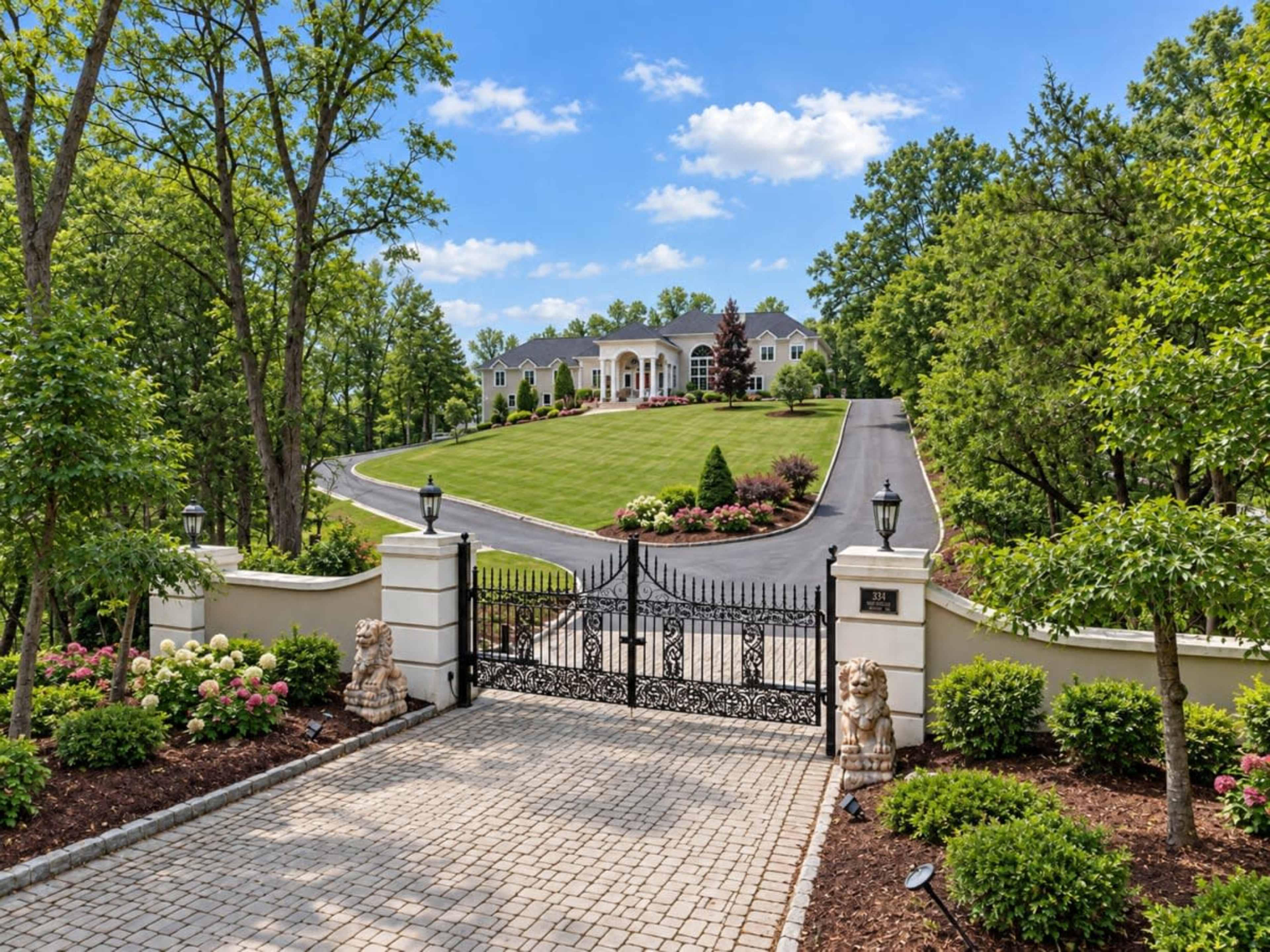 A grand entrance gated by ornamental wrought iron gates leads up a winding driveway to a large estate surrounded by lush greenery and landscaped gardens.