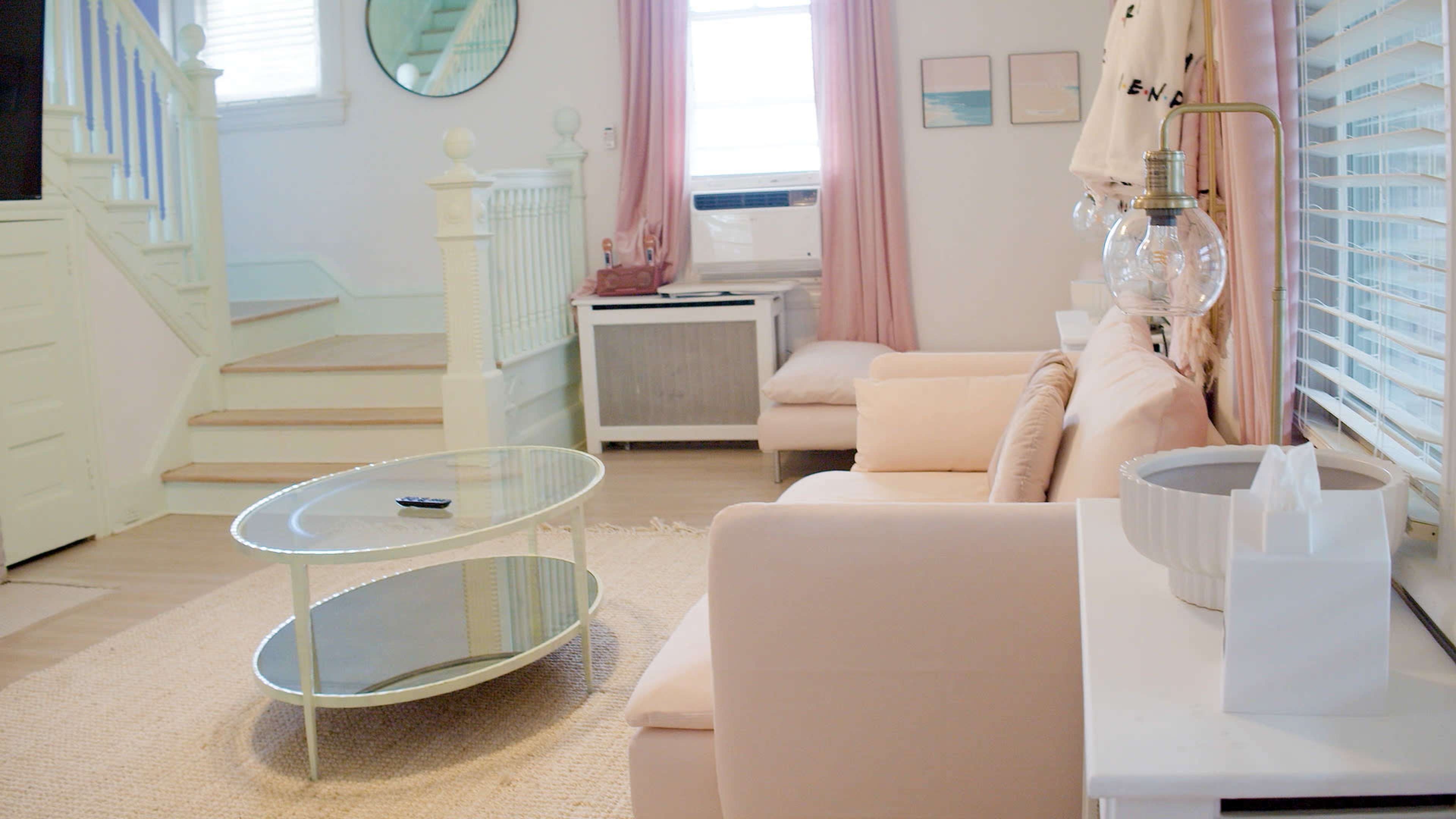 The image shows a cozy living room with a light-colored sofa, a circular glass coffee table, and soft pink curtains by a window.