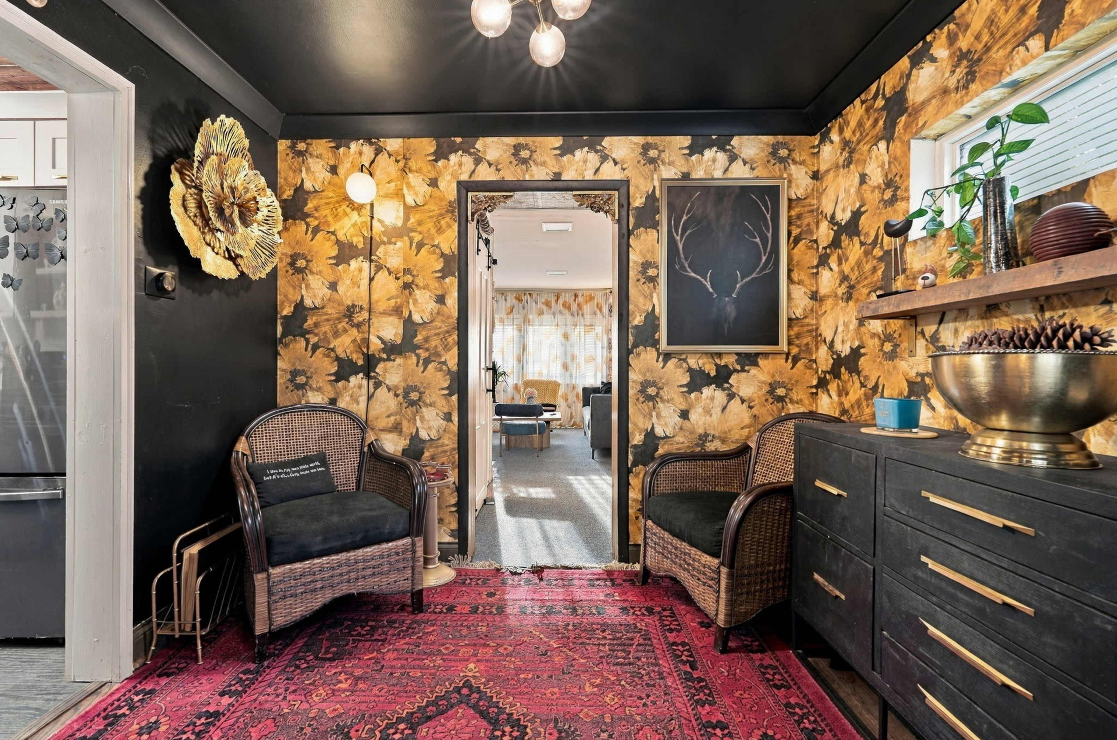 The image depicts a cozy entryway with floral wallpaper, a dark ceiling, two wicker chairs, and a bright area leading into another room with a rug and decorative elements.