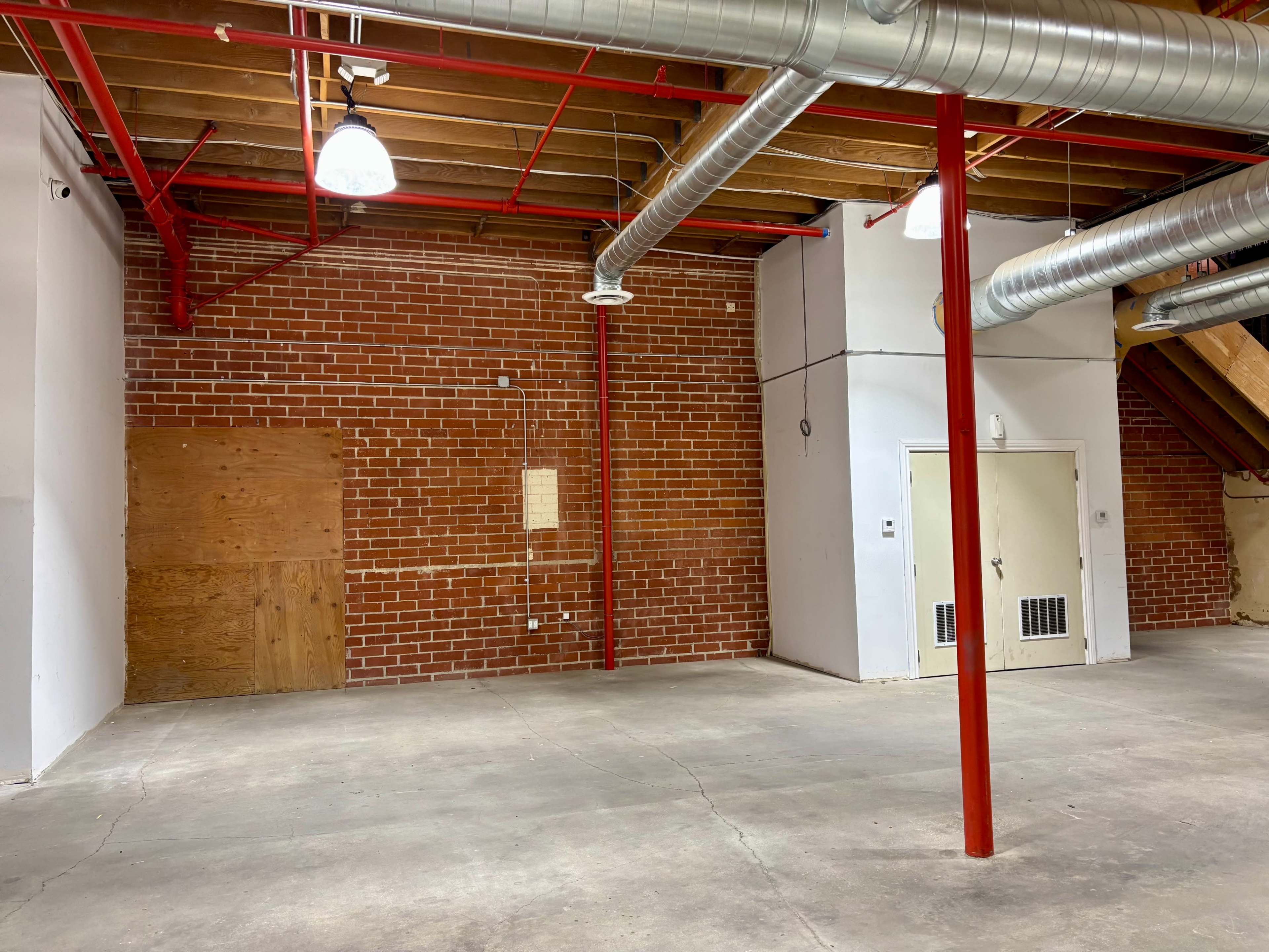 The image shows an empty industrial space with exposed brick walls, red piping, and overhead ductwork.