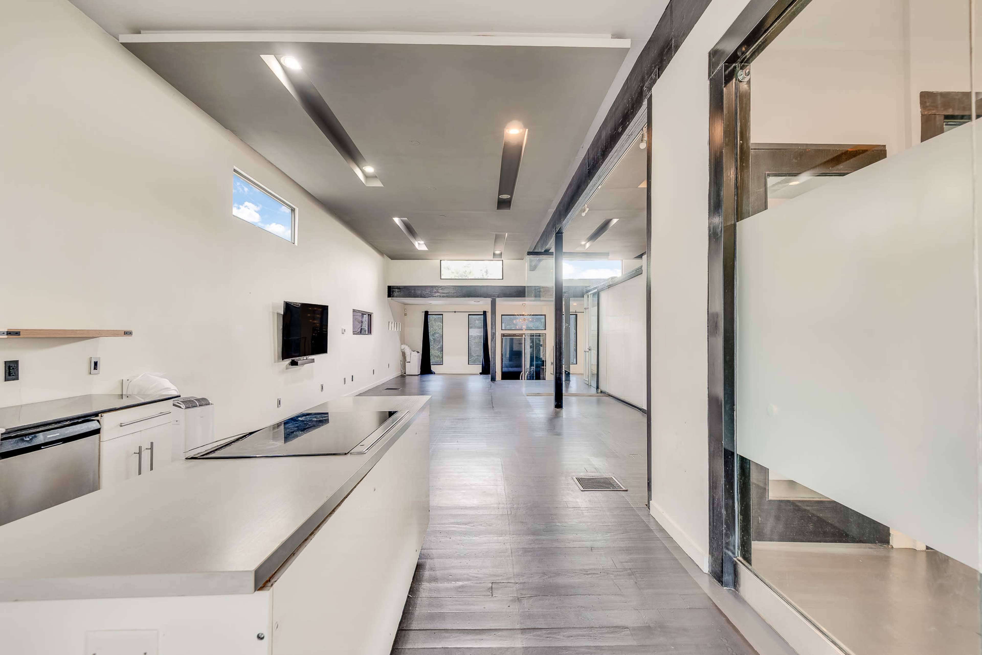 The image depicts a modern, open-plan interior featuring a kitchen area on the left and a long corridor leading to a glass door at the far end.