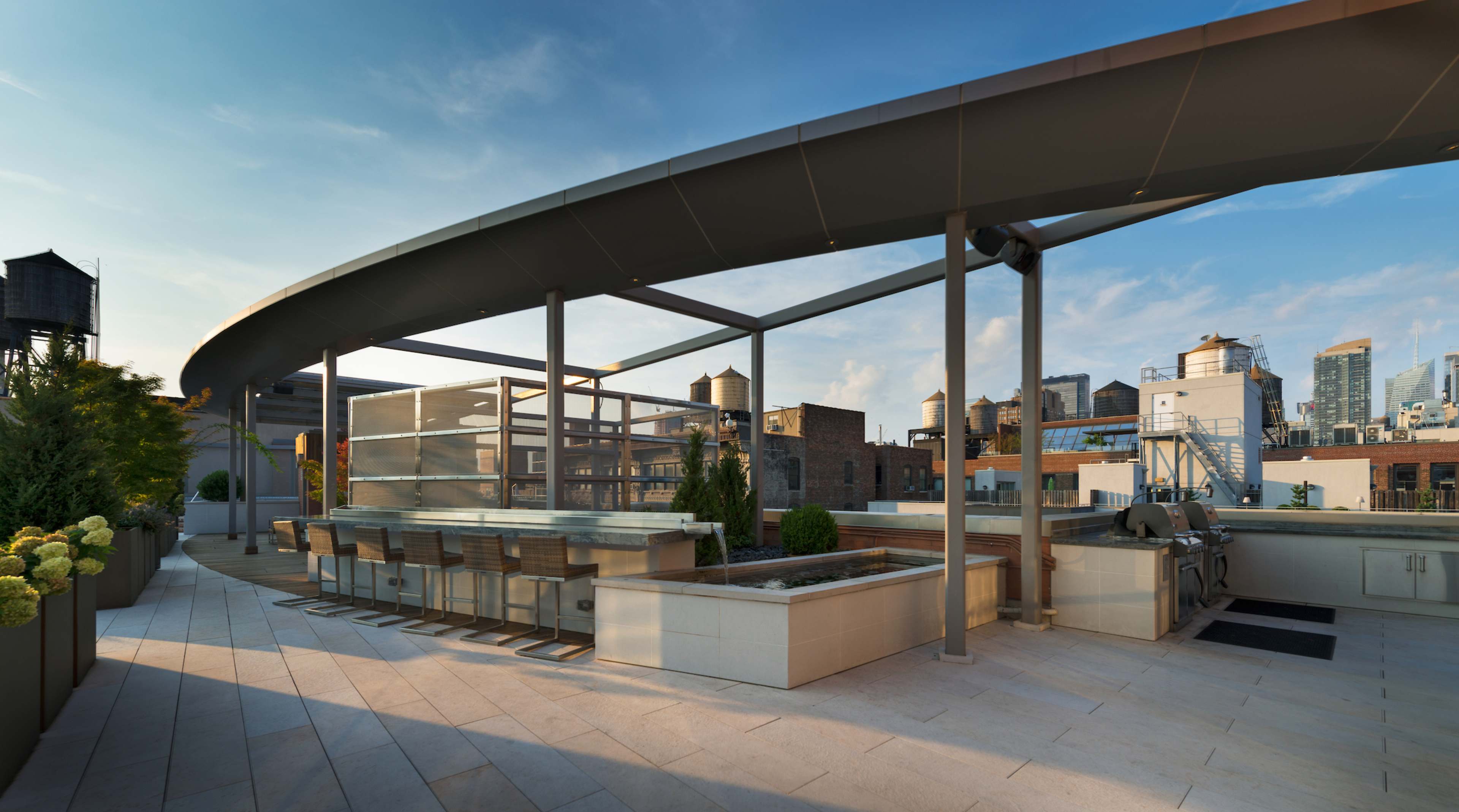 The image shows a modern rooftop terrace with seating, greenery, and a skyline view of city buildings.