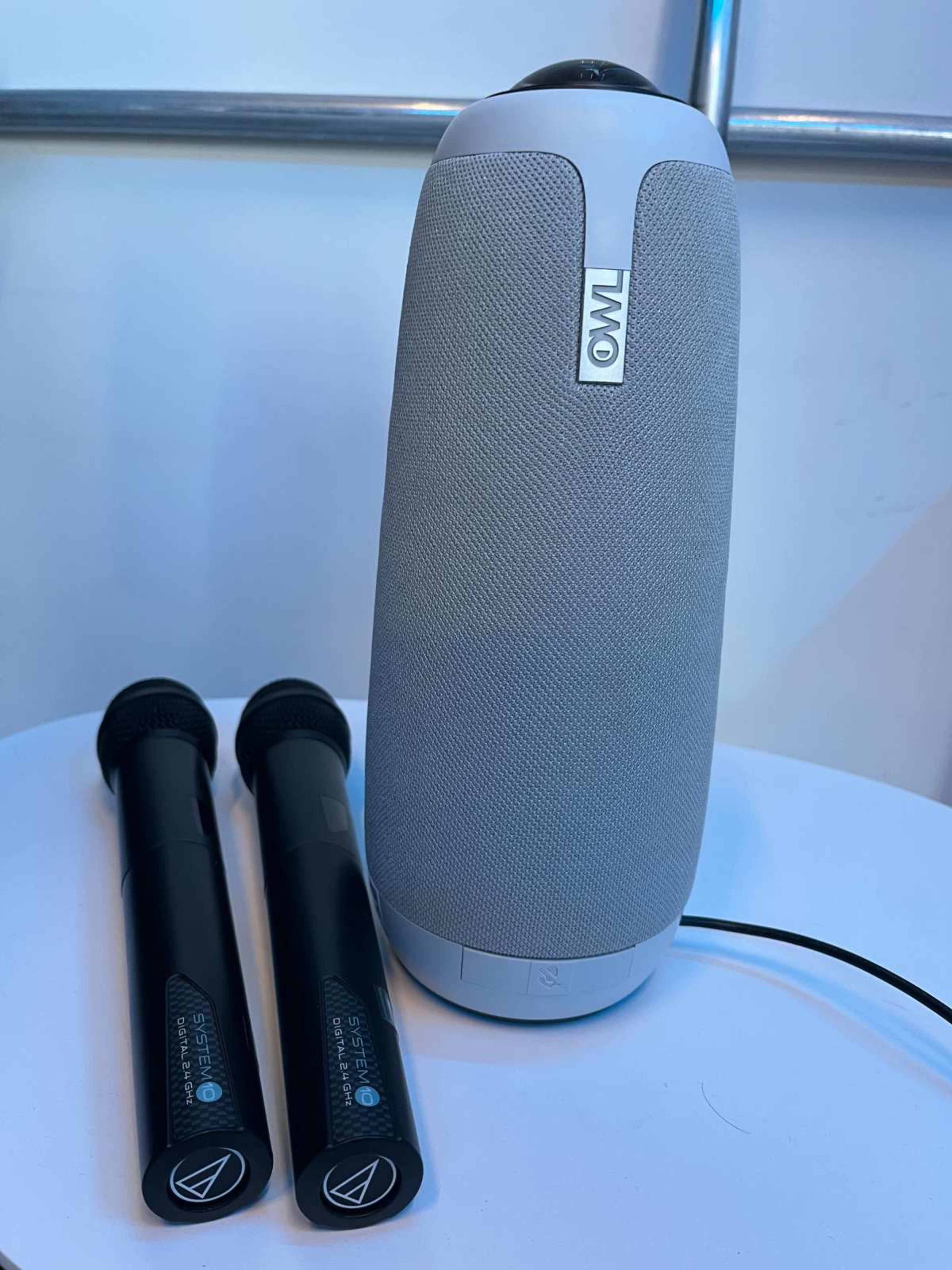 The image shows a portable speaker with a grey fabric exterior and two black microphones placed beside it on a white table.