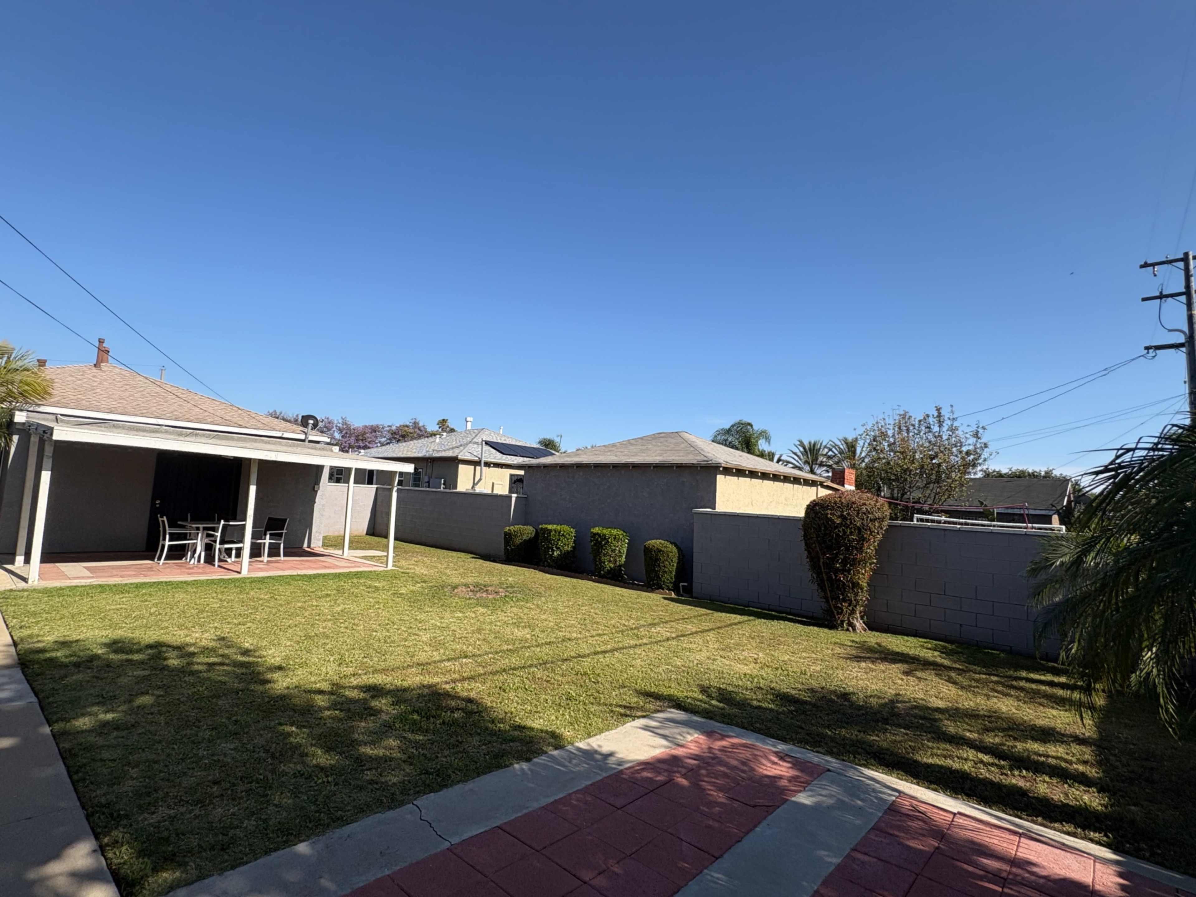 The image shows a backyard with a grassy area, a covered patio, and several shrubs along a fence under a clear blue sky.