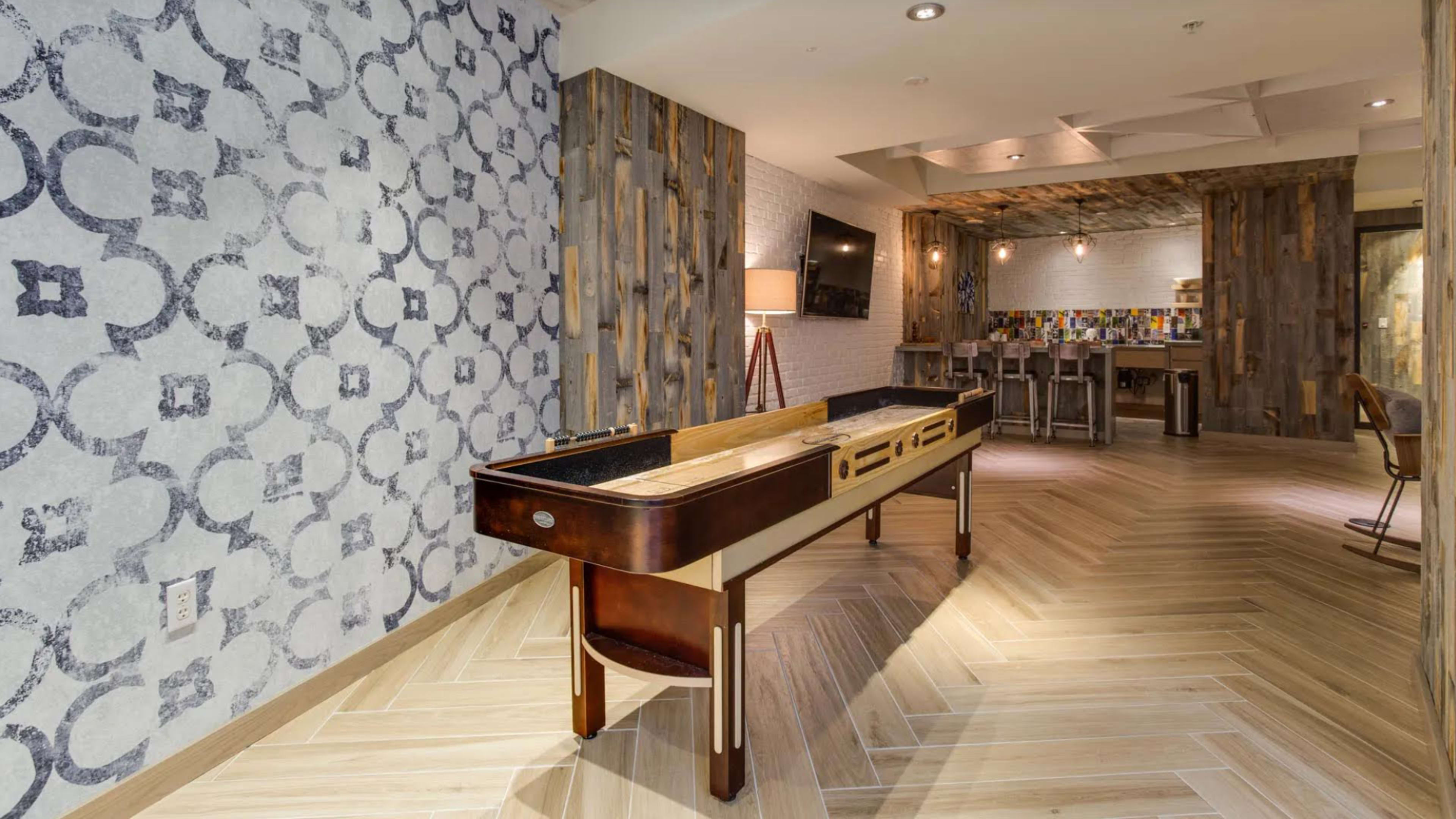 A game room with a shuffleboard table, patterned wallpaper, and a casual seating area in the background.