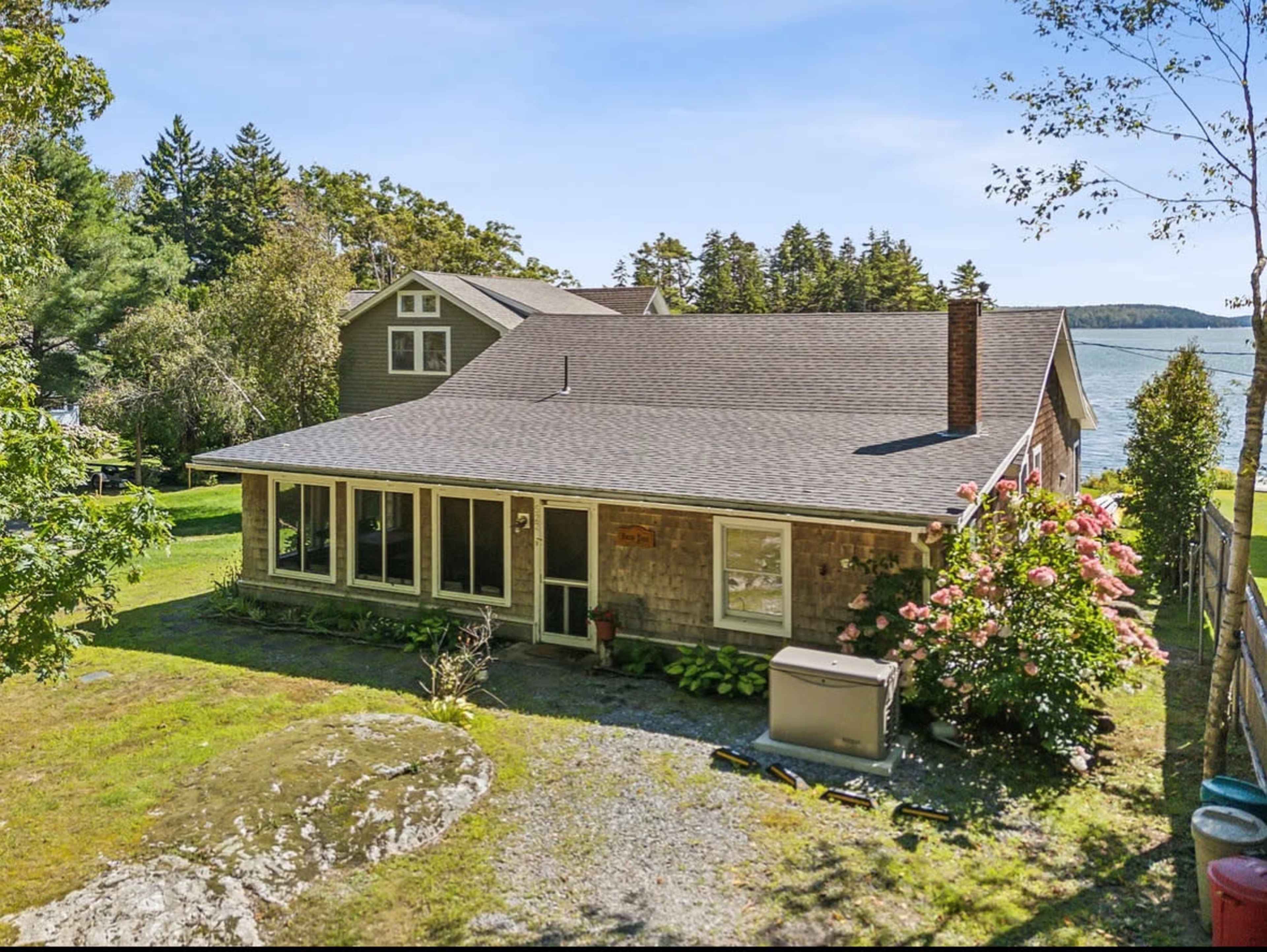 The image shows a single-story house with a stone exterior, surrounded by greenery and trees, with a view of water in the background.