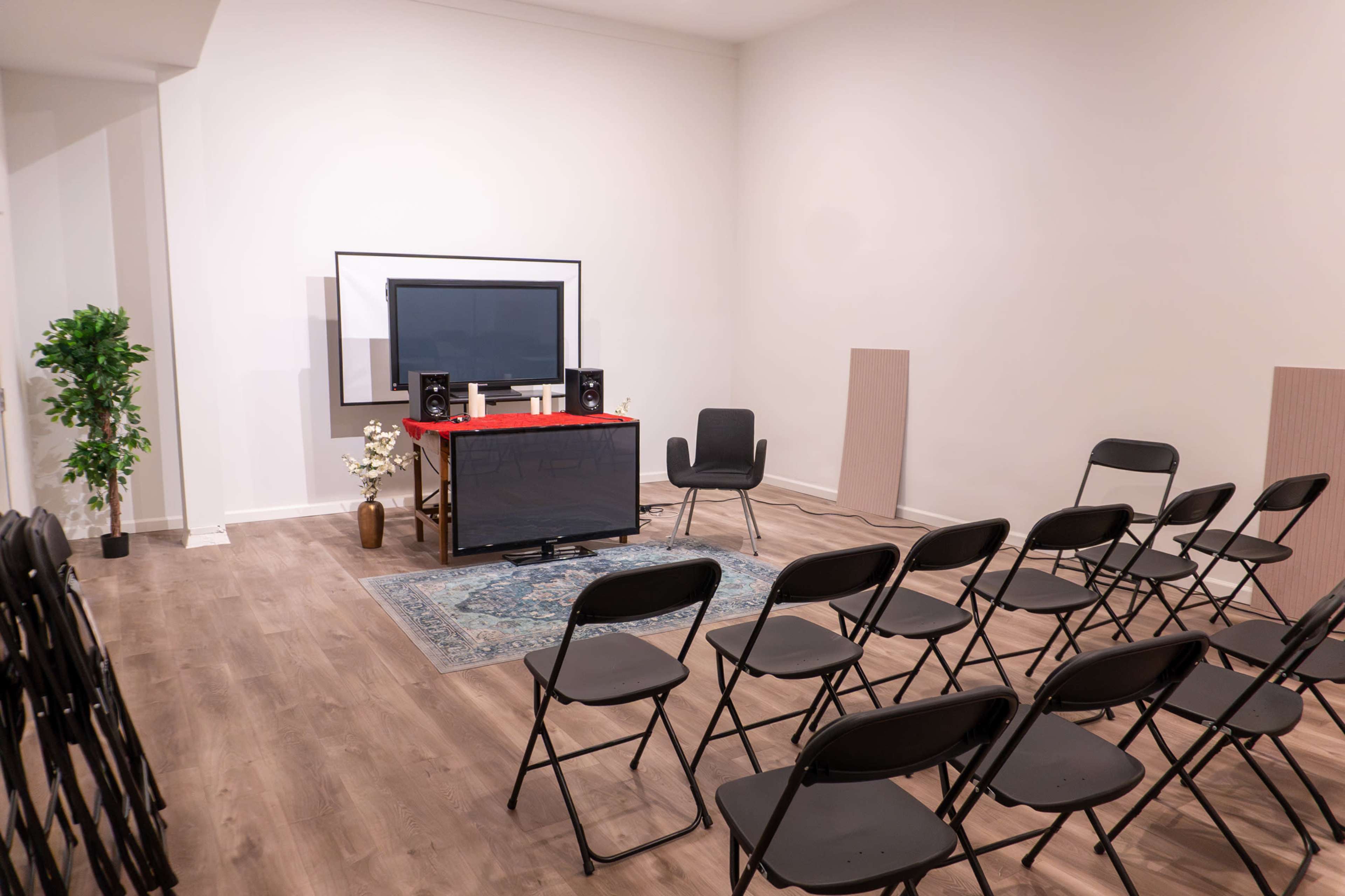 A small, sparsely furnished room contains rows of black folding chairs facing a television and sound system on a low table, with a decorative rug on the floor.