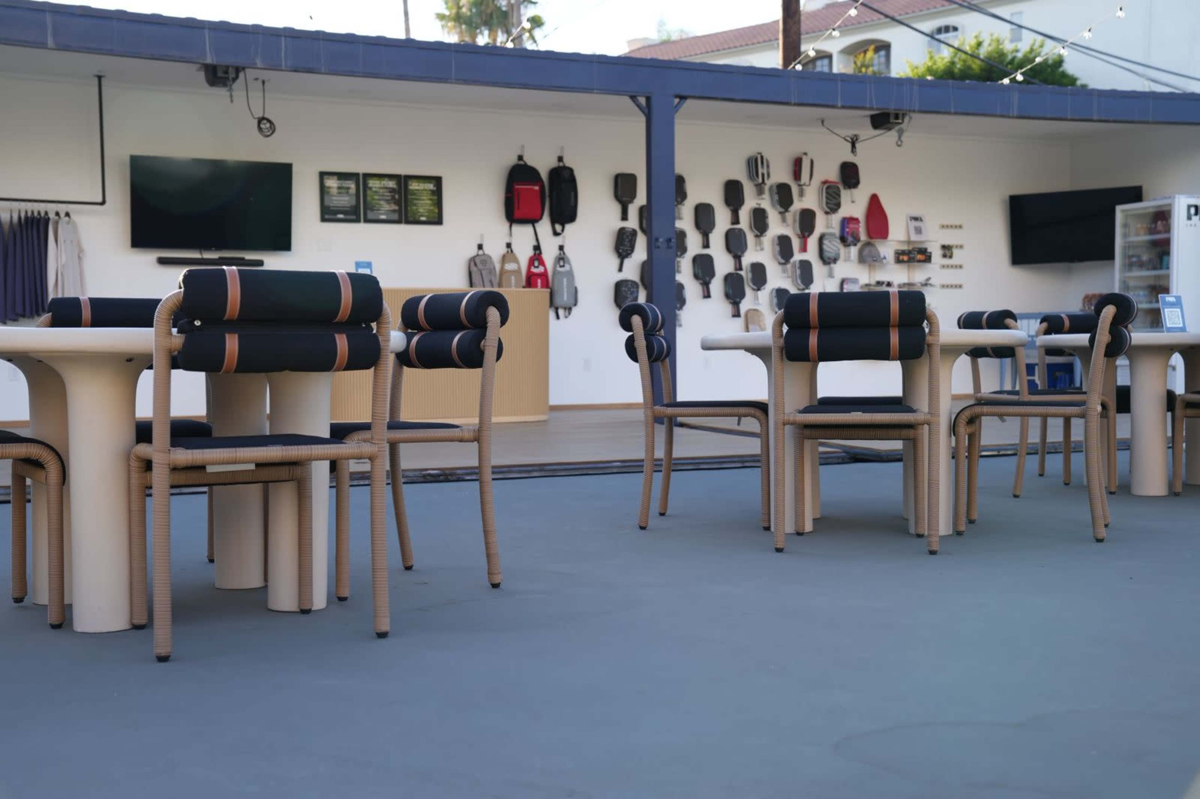 The image shows a patio area with several tables and chairs, surrounded by a wall displaying various bags and equipment.