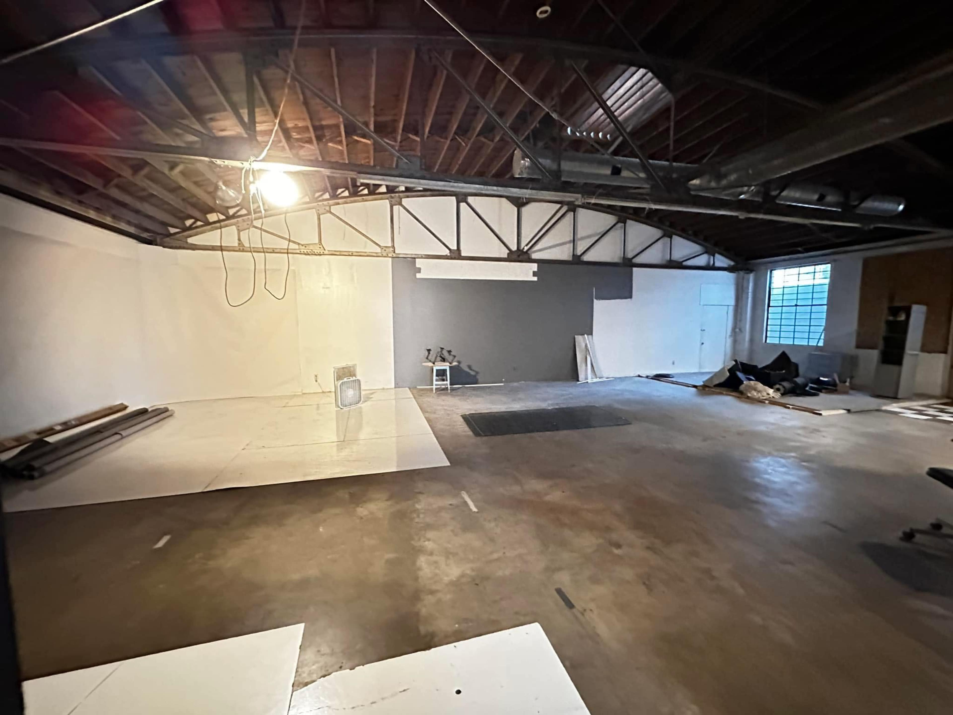 The image shows an empty industrial-style room with exposed beams, concrete flooring, and some materials scattered along the walls.