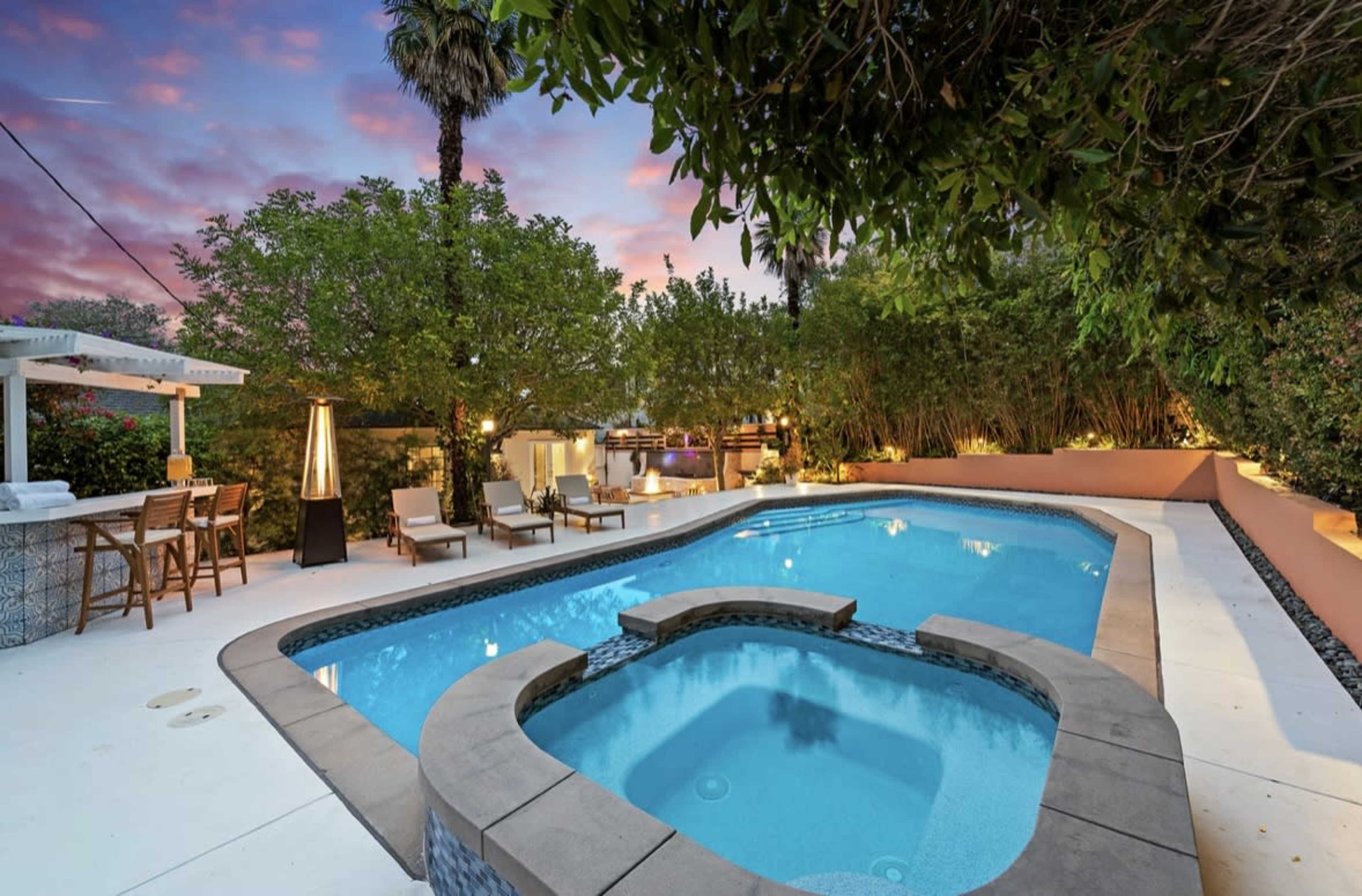A tranquil backyard features a rectangular swimming pool with a spa, surrounded by lounge chairs and lush greenery under a sunset sky.