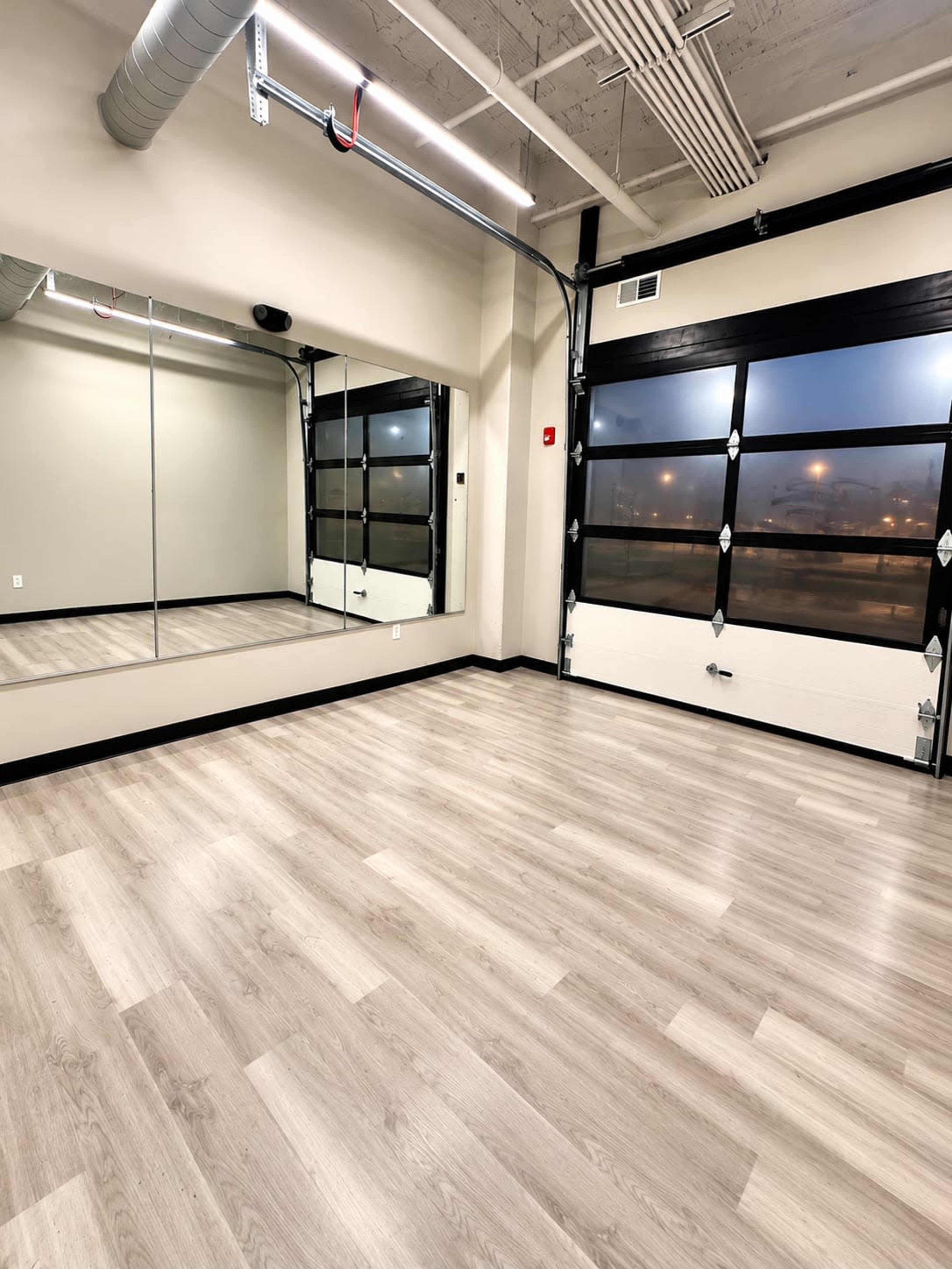 The image shows an empty indoor space with light-colored laminate flooring, large mirrors on one wall, and a garage door partially open, allowing natural light to filter in.
