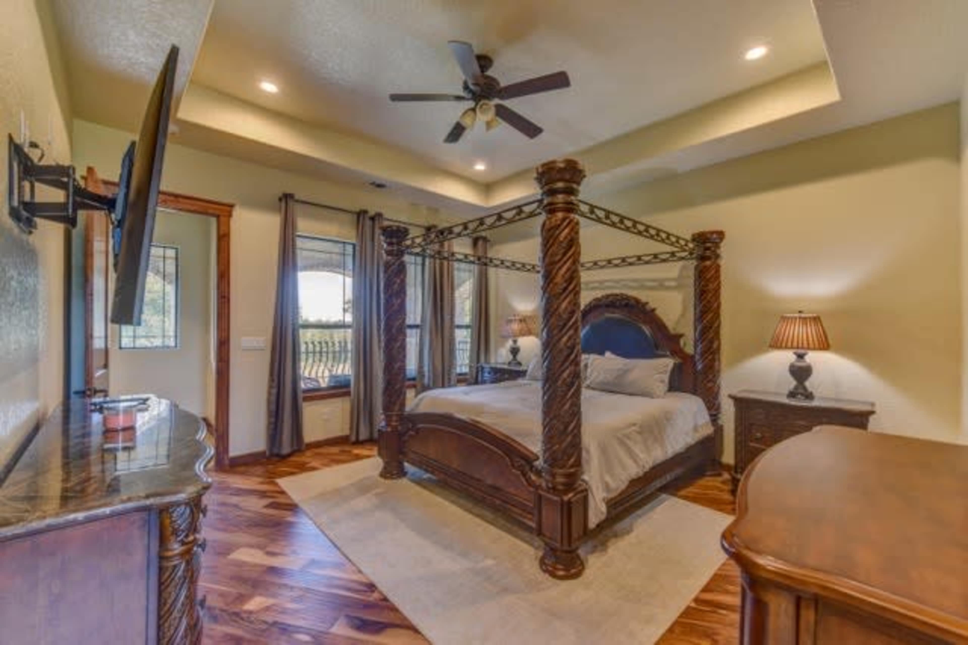 A spacious bedroom featuring a four-poster bed, wooden furniture, a television mounted on the wall, and large windows with natural light.