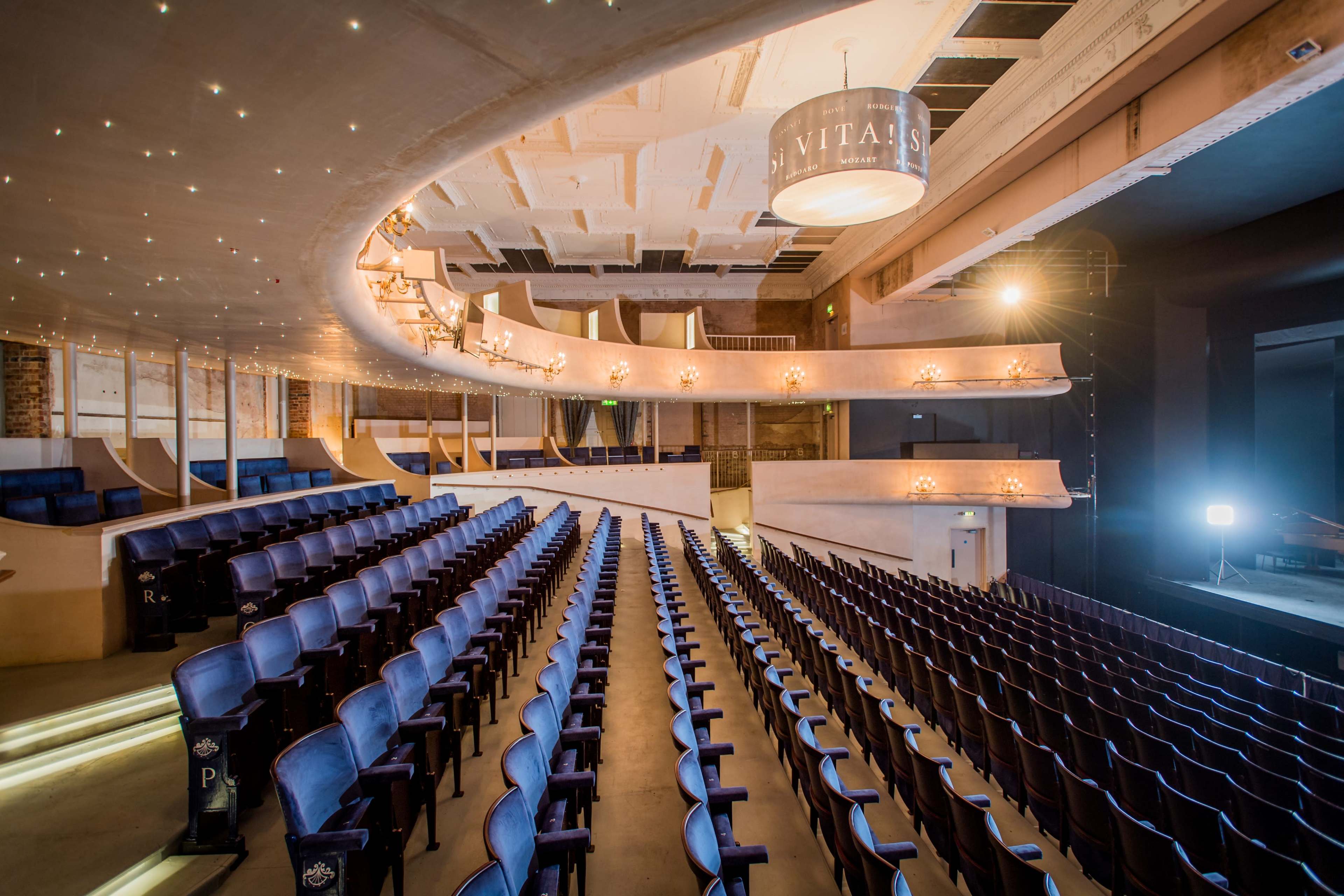 The image shows a theater interior with rows of blue seats and a balcony overlooking a stage, illuminated by soft overhead lighting.