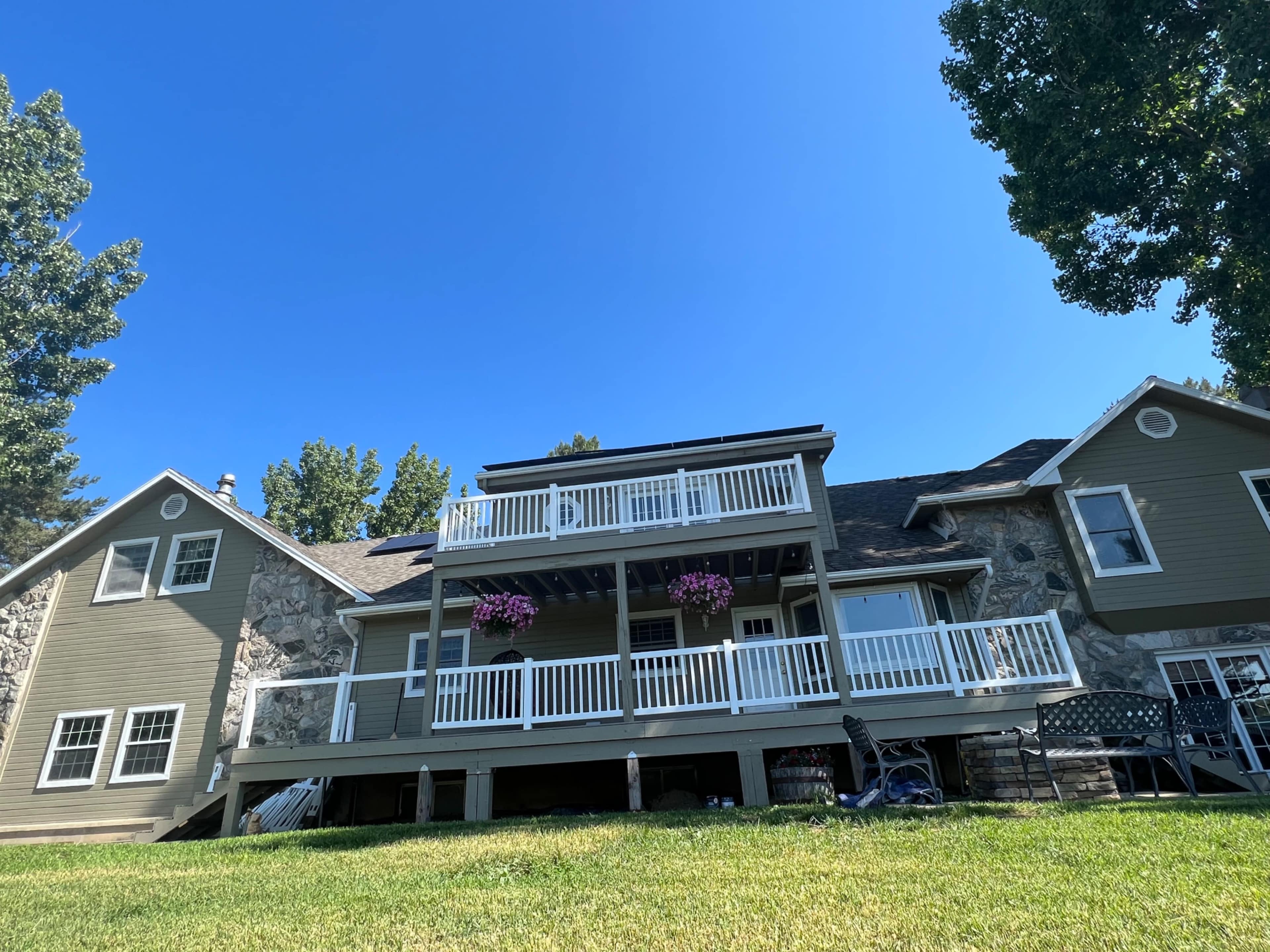 A two-story house with a stone facade features a large deck adorned with hanging flower baskets, situated on a grassy lawn under a clear blue sky.