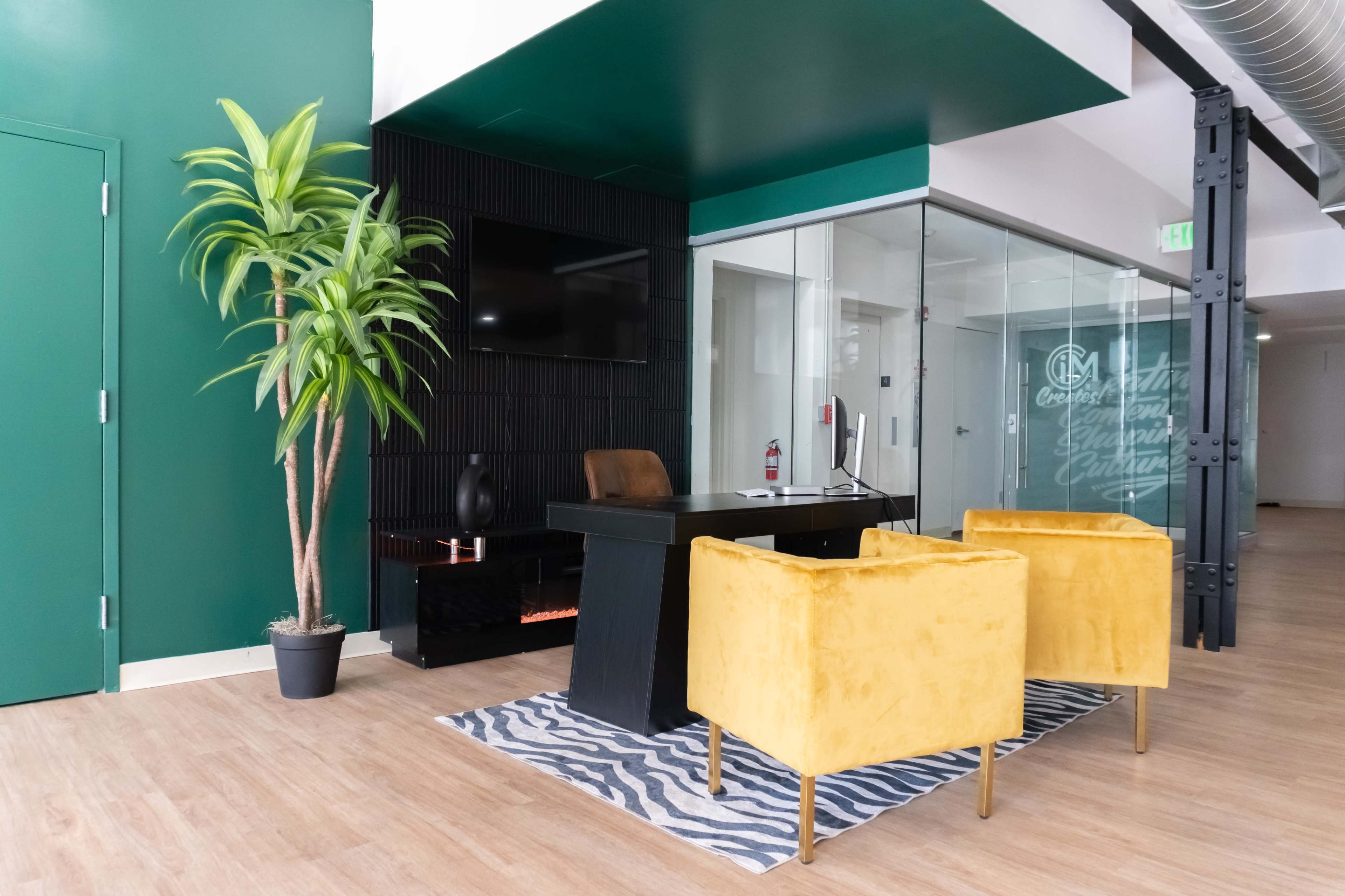 The image shows a modern office reception area featuring a black desk, two yellow chairs, and a potted plant against a green and white wall.