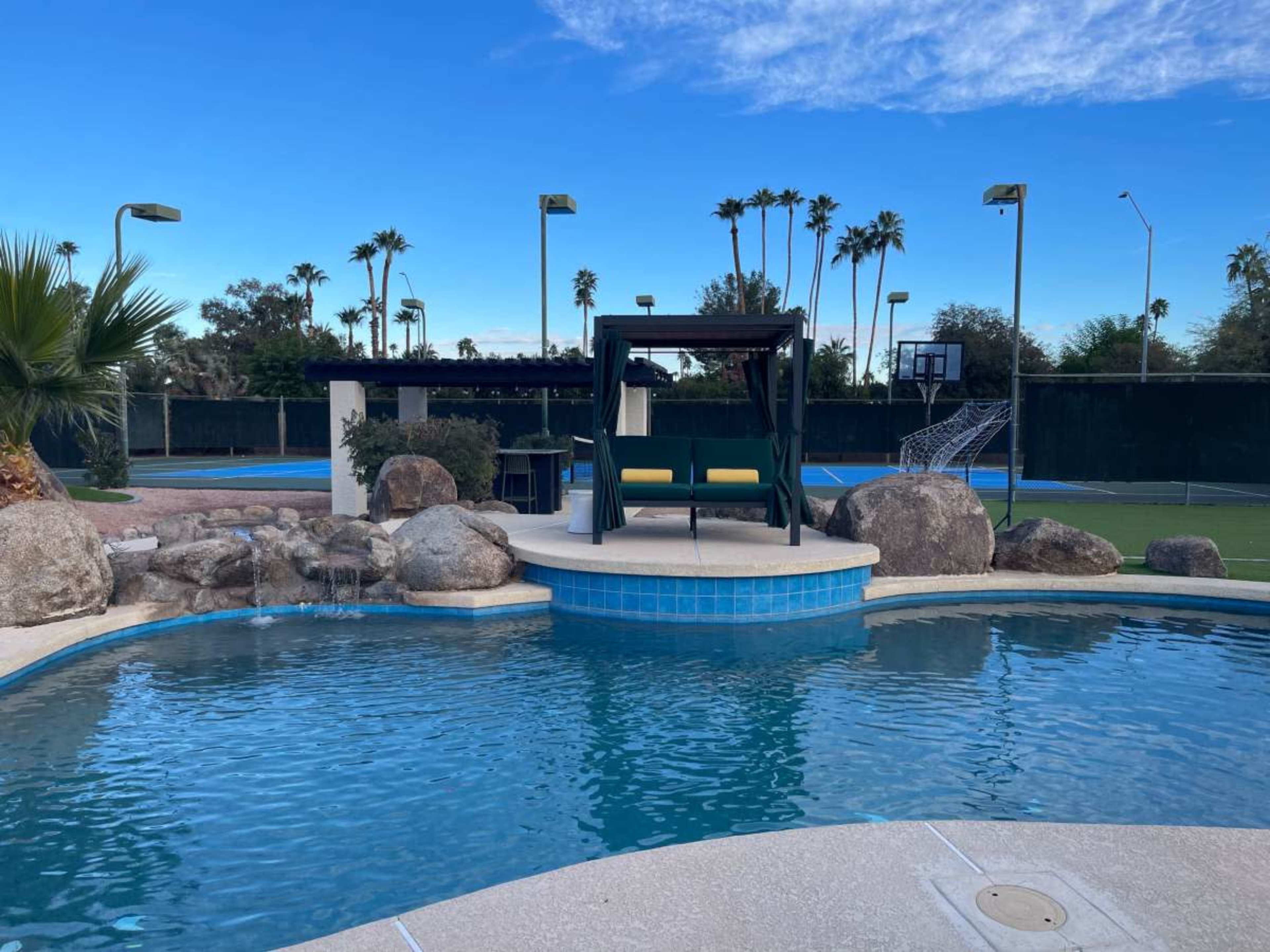 A swimming pool surrounded by rocks and palm trees features a shaded seating area and a basketball court in the background.