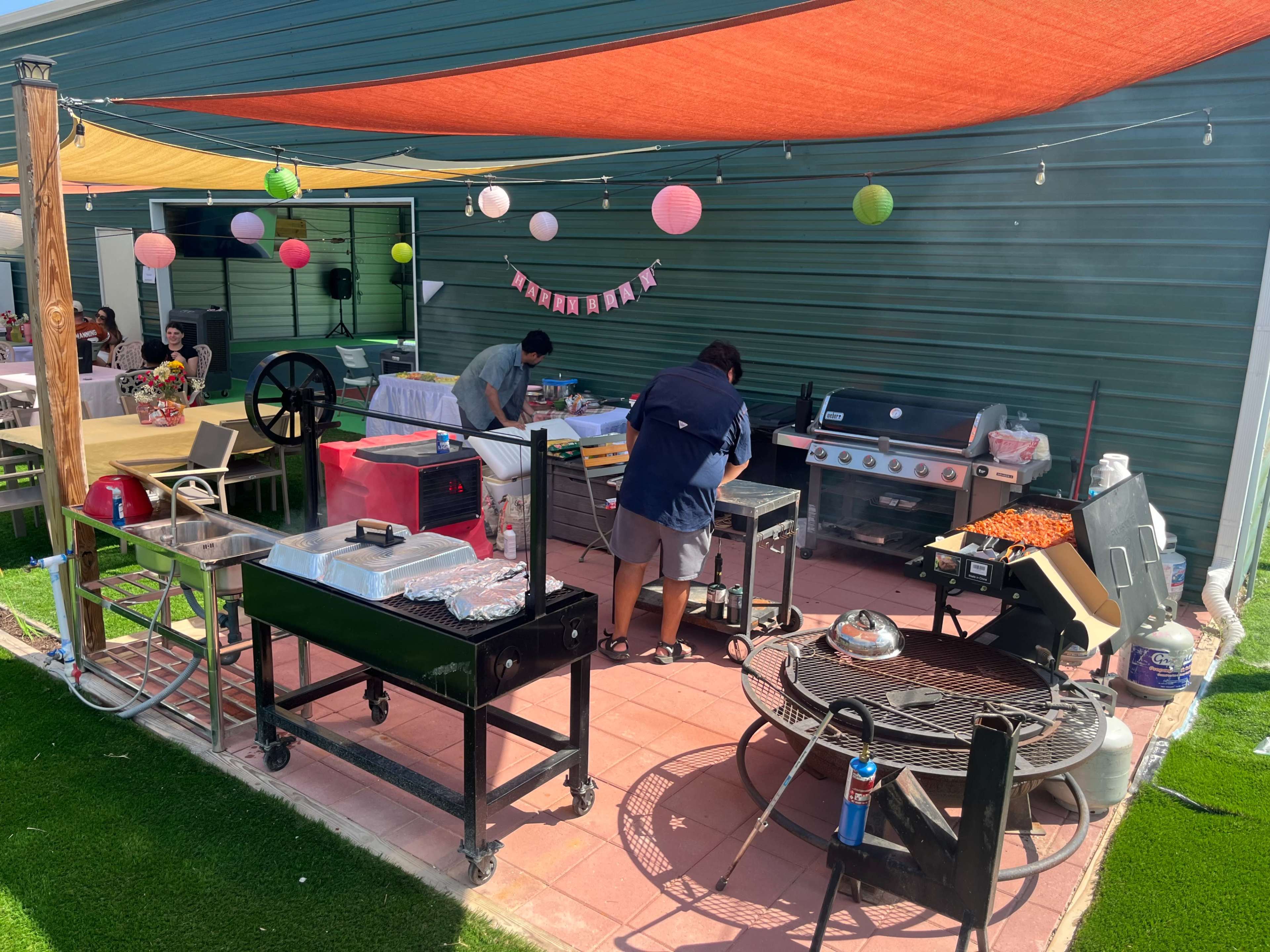 Two people are preparing food on a grill in a lively outdoor kitchen area, decorated with colorful lanterns and shaded by vibrant awnings.