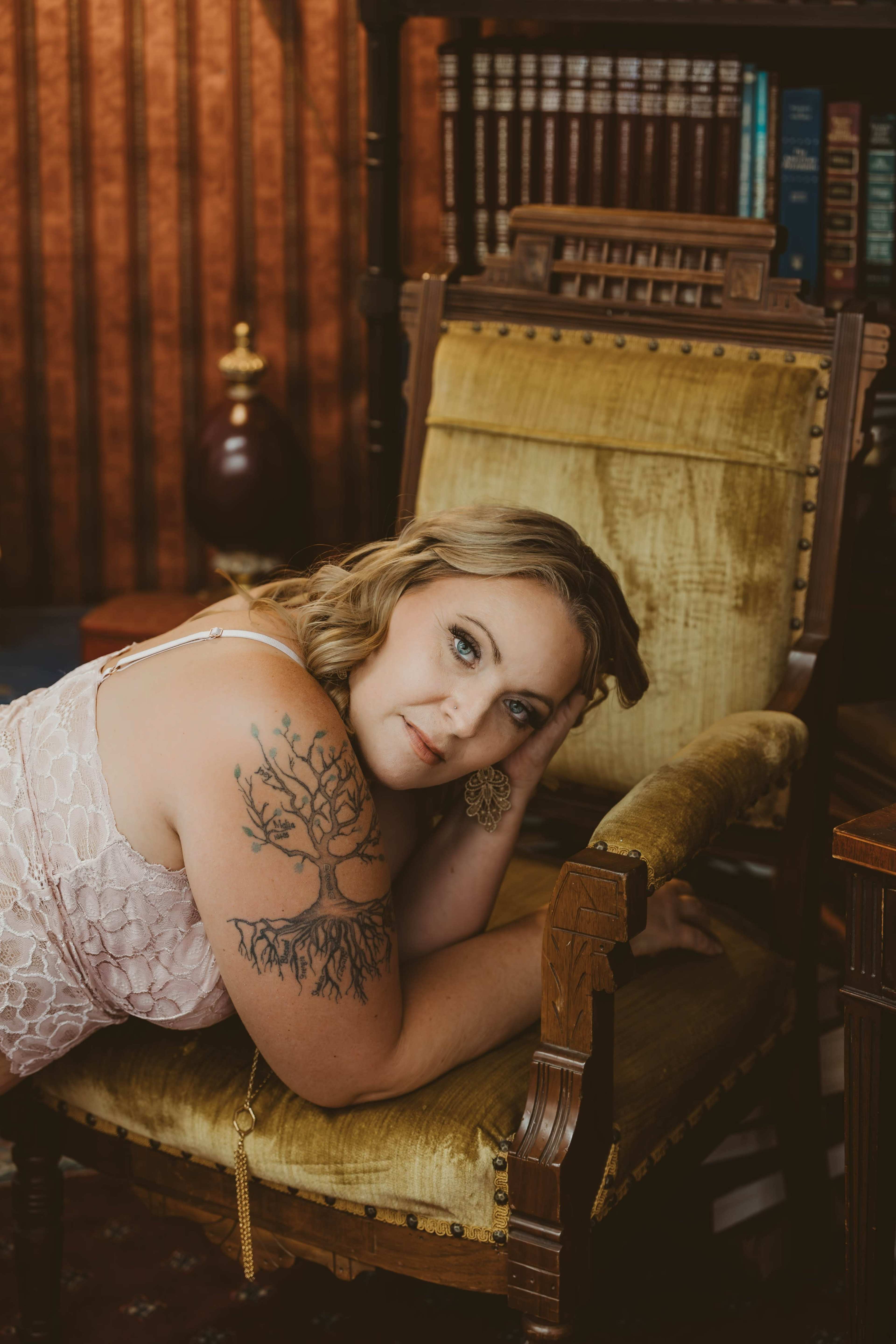 A woman with long hair rests her head on her hand while lying on an antique chair in a warmly lit room filled with books.
