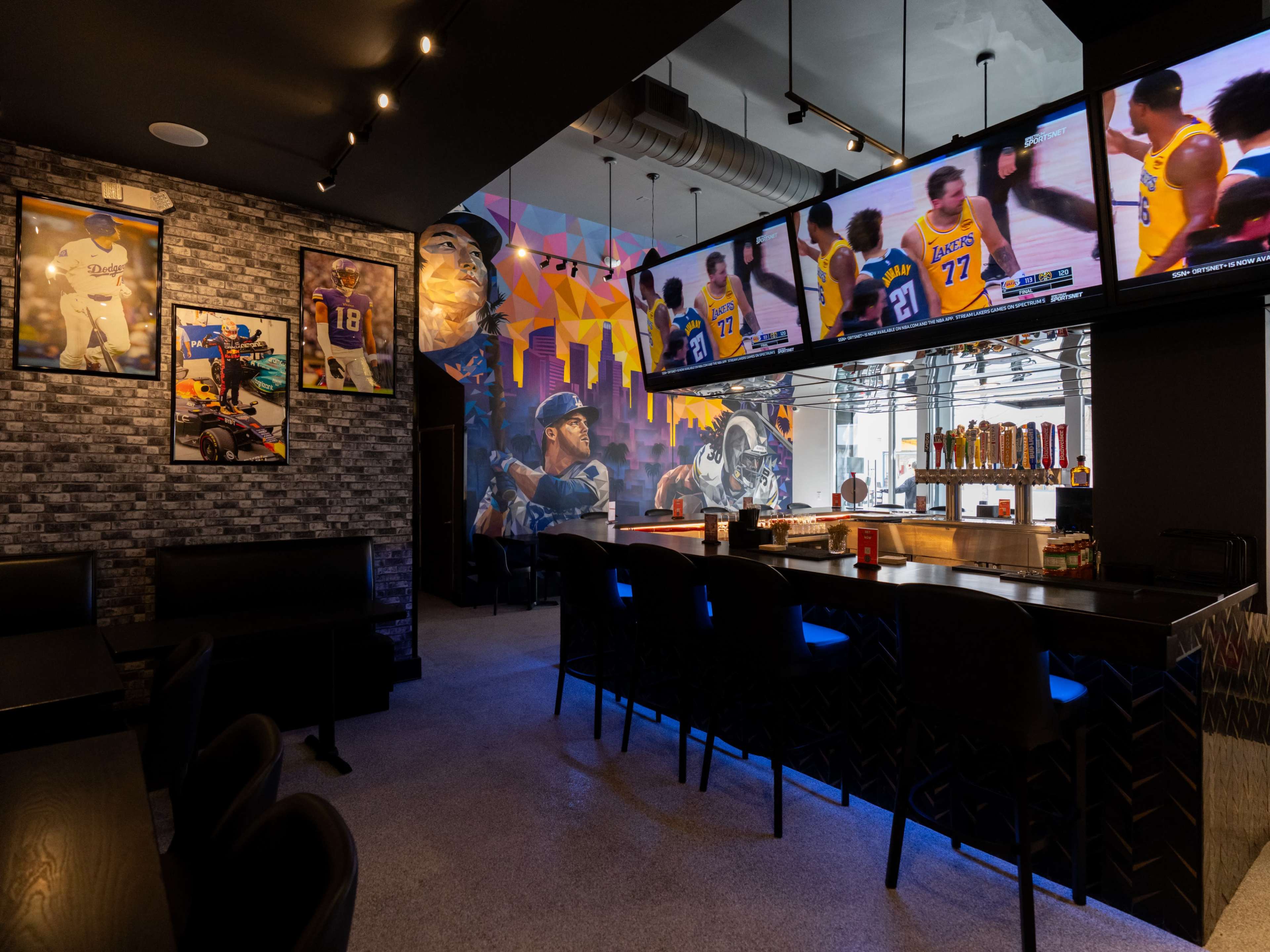 The image shows a modern sports bar featuring large television screens showing a basketball game, a stylish bar area, and walls adorned with sports-themed artwork and framed photographs.