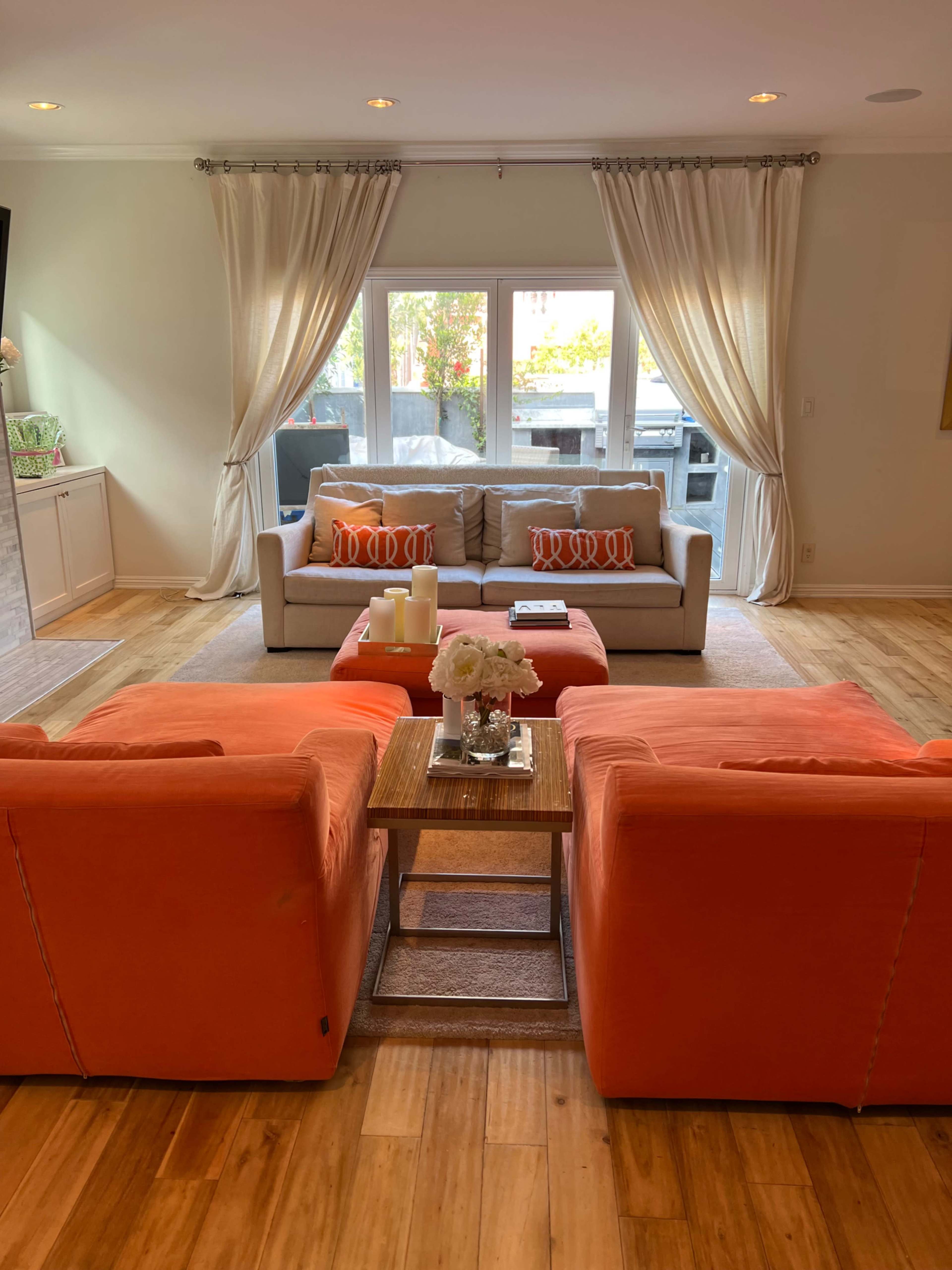 The living room features a light gray sofa with orange and white pillows, flanked by two orange armchairs, centered around a wooden coffee table with a vase of flowers.