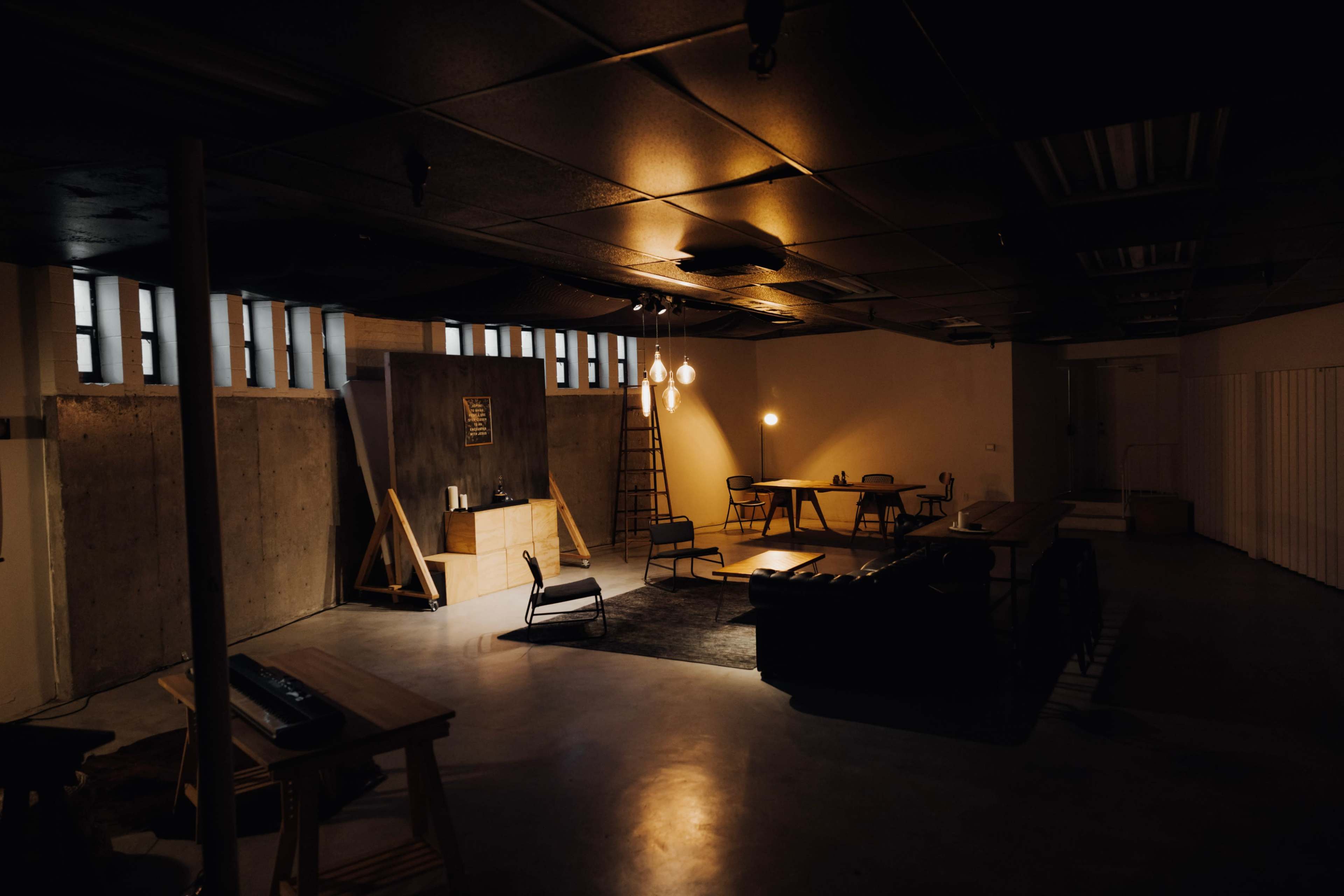 The image shows a dimly lit modern interior space featuring concrete walls, scattered furniture, and hanging light fixtures.