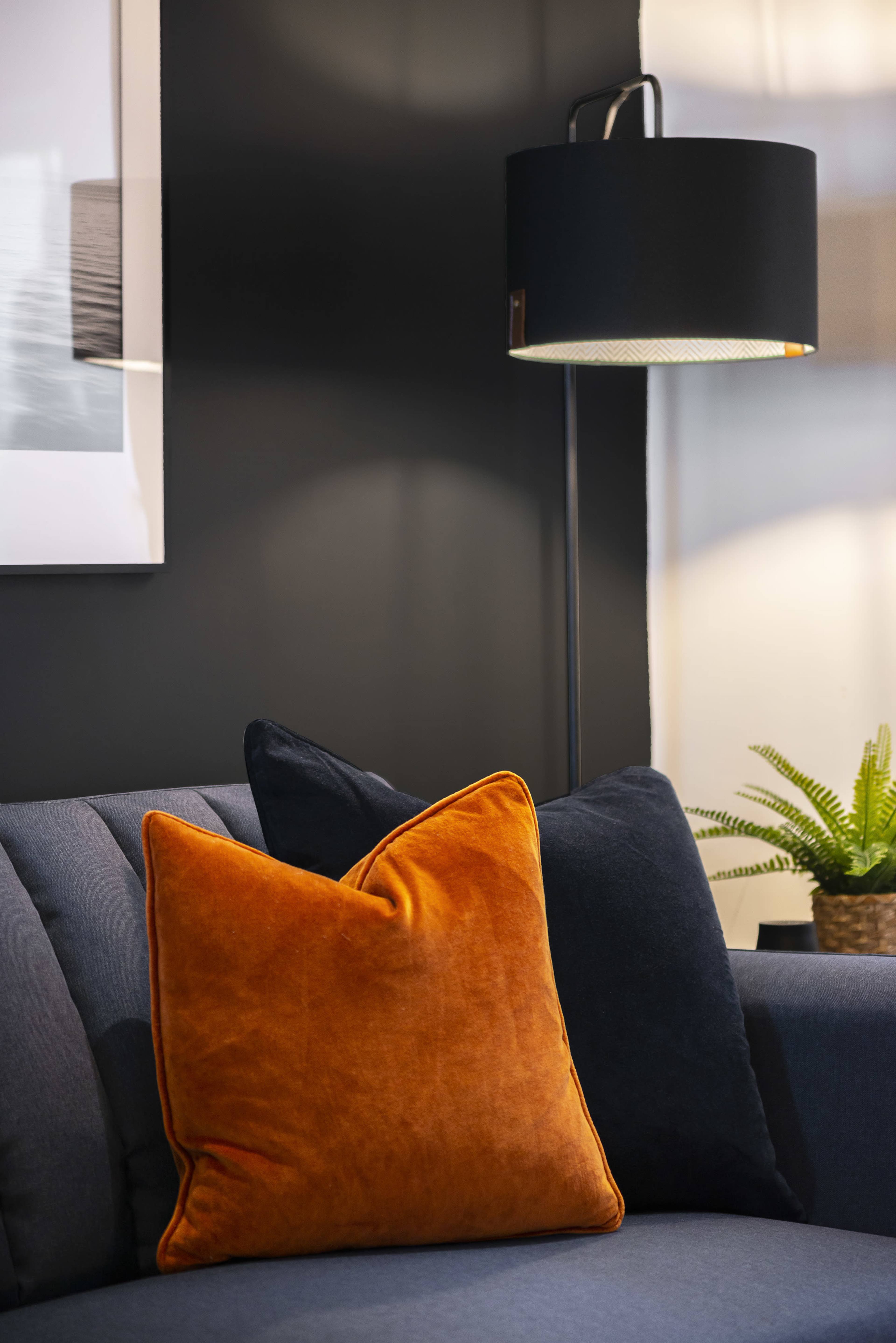 A dark wall behind a couch with two decorative pillows and a standing lamp next to a small plant.