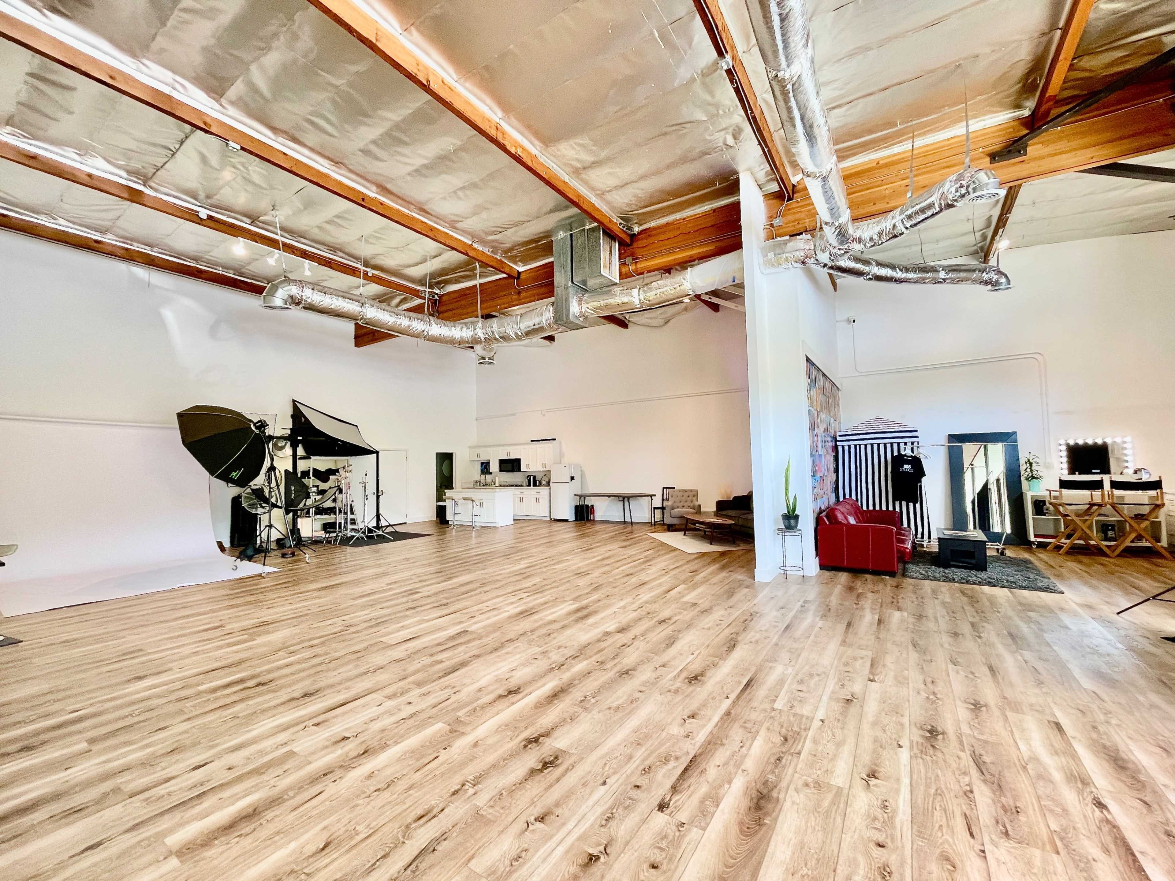 An expansive, well-lit studio space with wooden flooring, industrial-style ceiling beams, and various photography equipment positioned along one side.