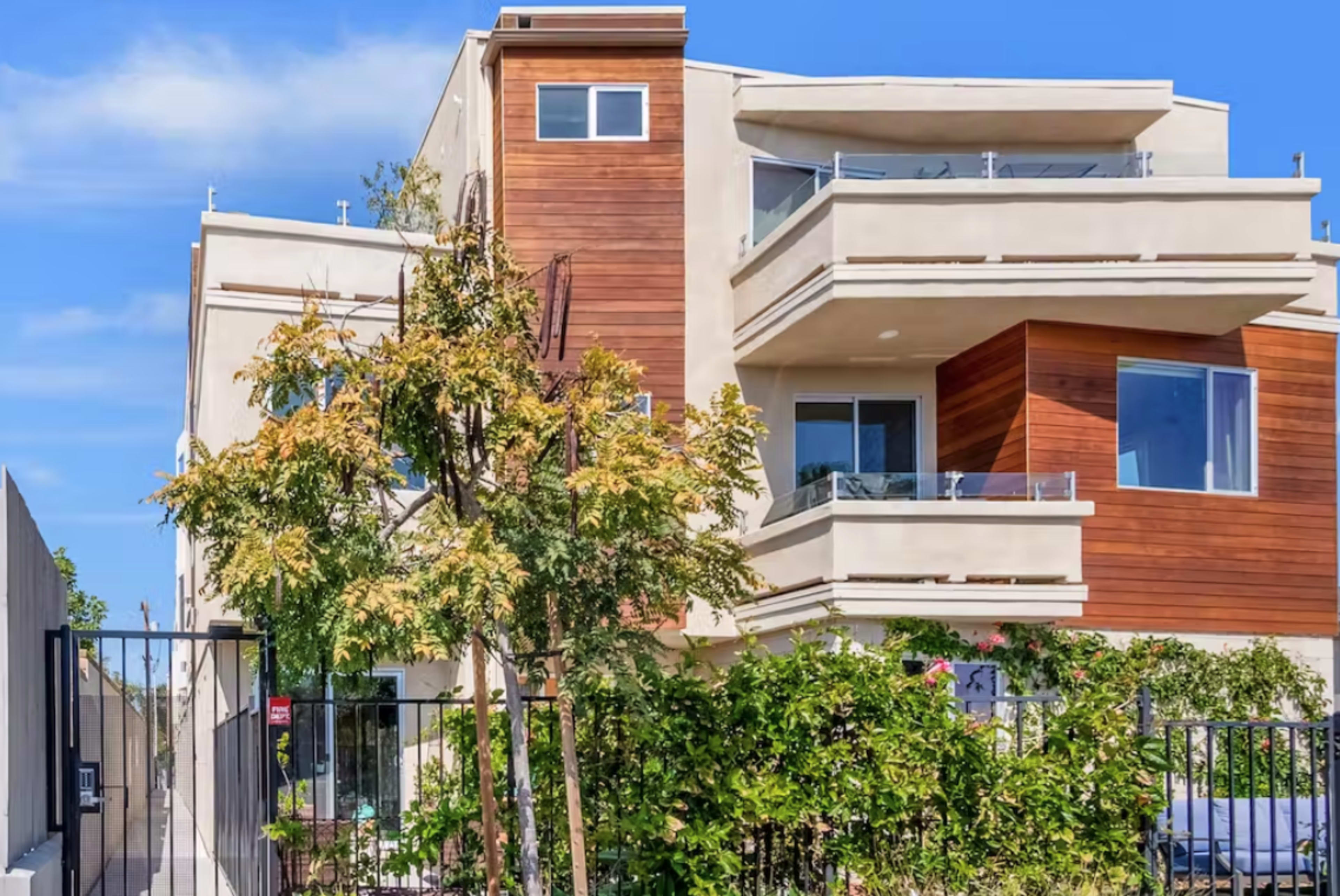 A modern three-story building features a combination of stucco and wooden paneling, with balconies and surrounding greenery.