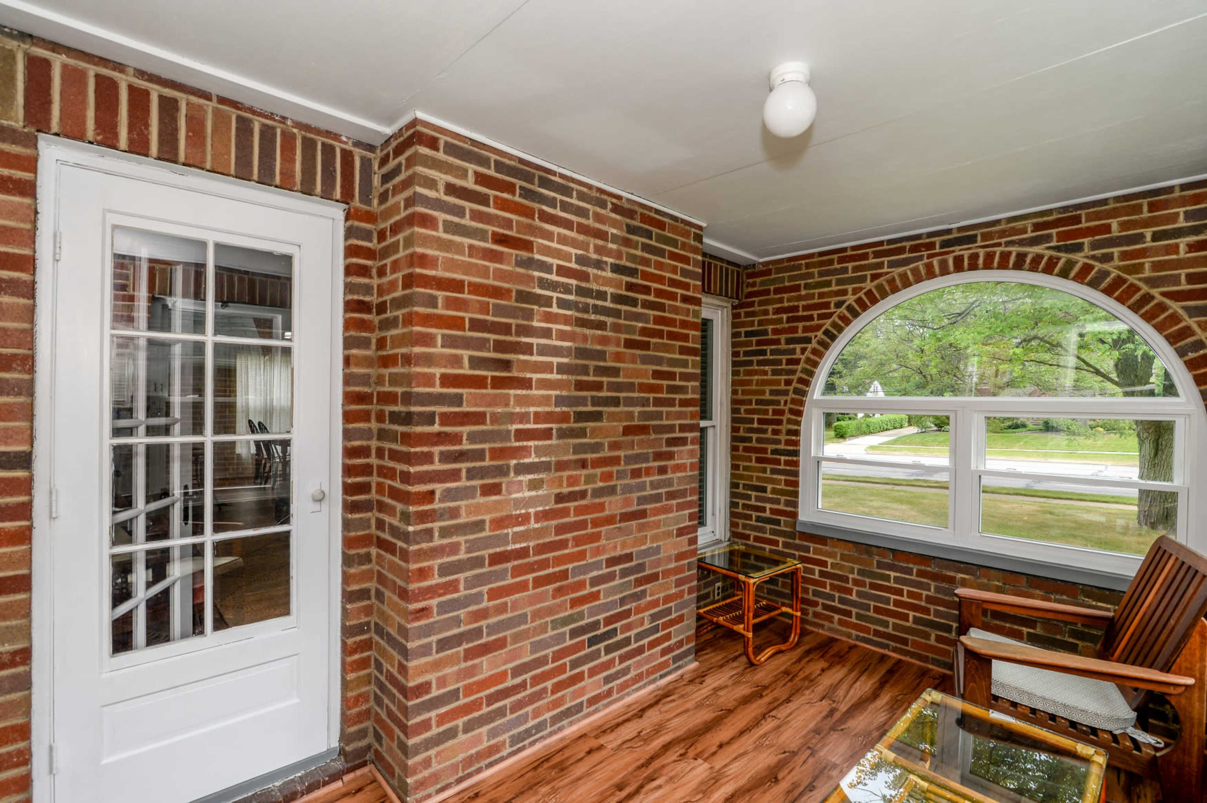 A porch with brick walls, wooden flooring, a round window, a door, and two chairs.