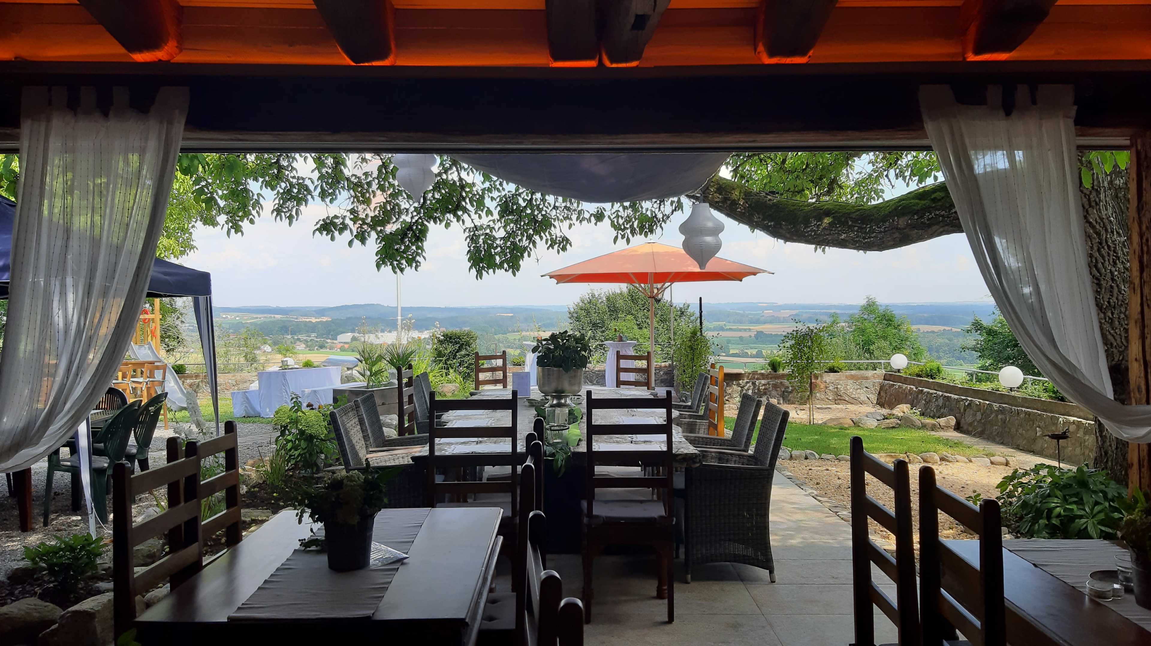 The image shows a restaurant setting with wooden tables and chairs arranged under a canopy, offering a view of a landscape with an umbrella and greenery in the background.