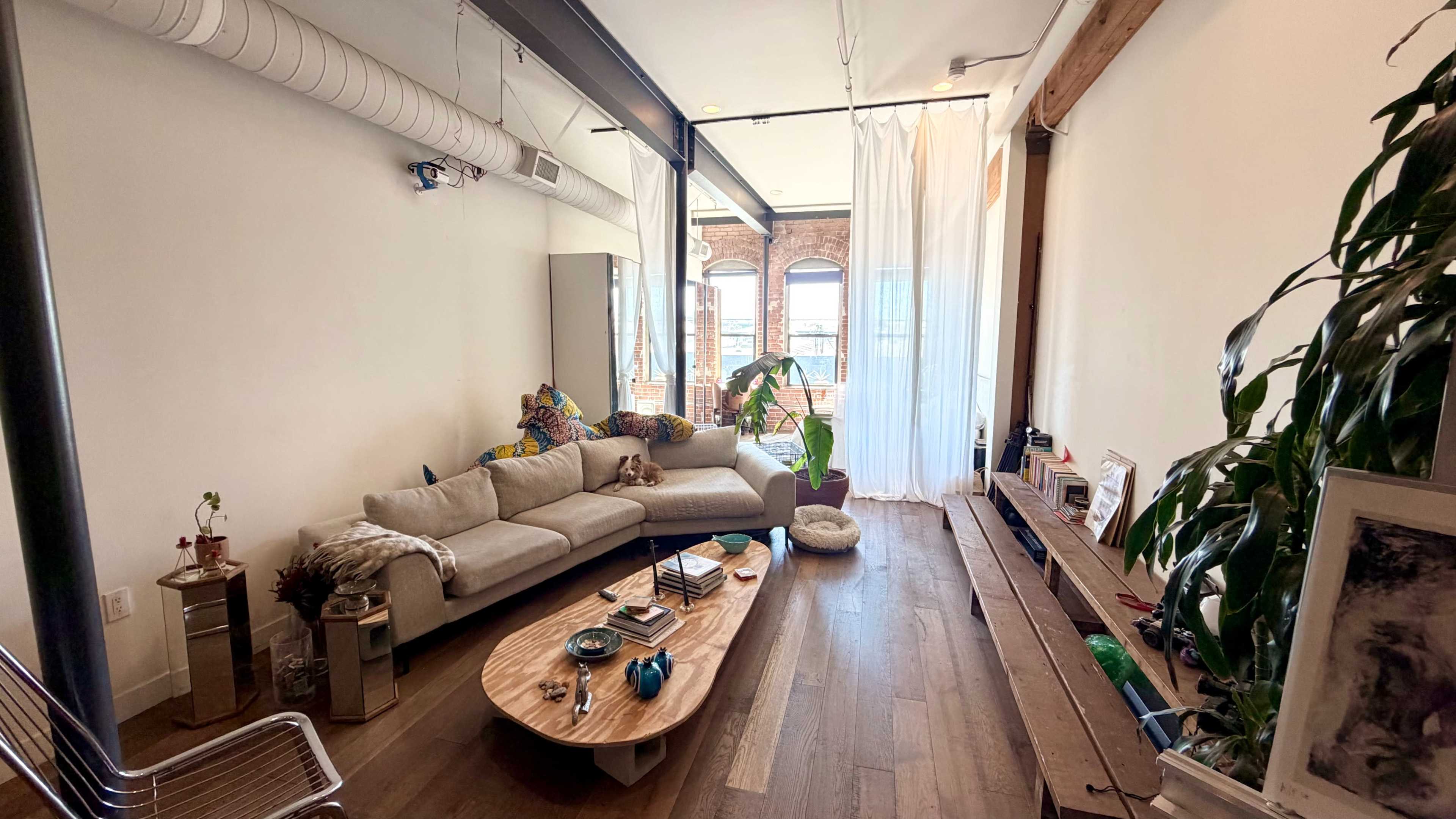 The image shows a cozy living room with a light-colored sofa, a wooden coffee table, and large windows that provide a view of a bright outdoor space.