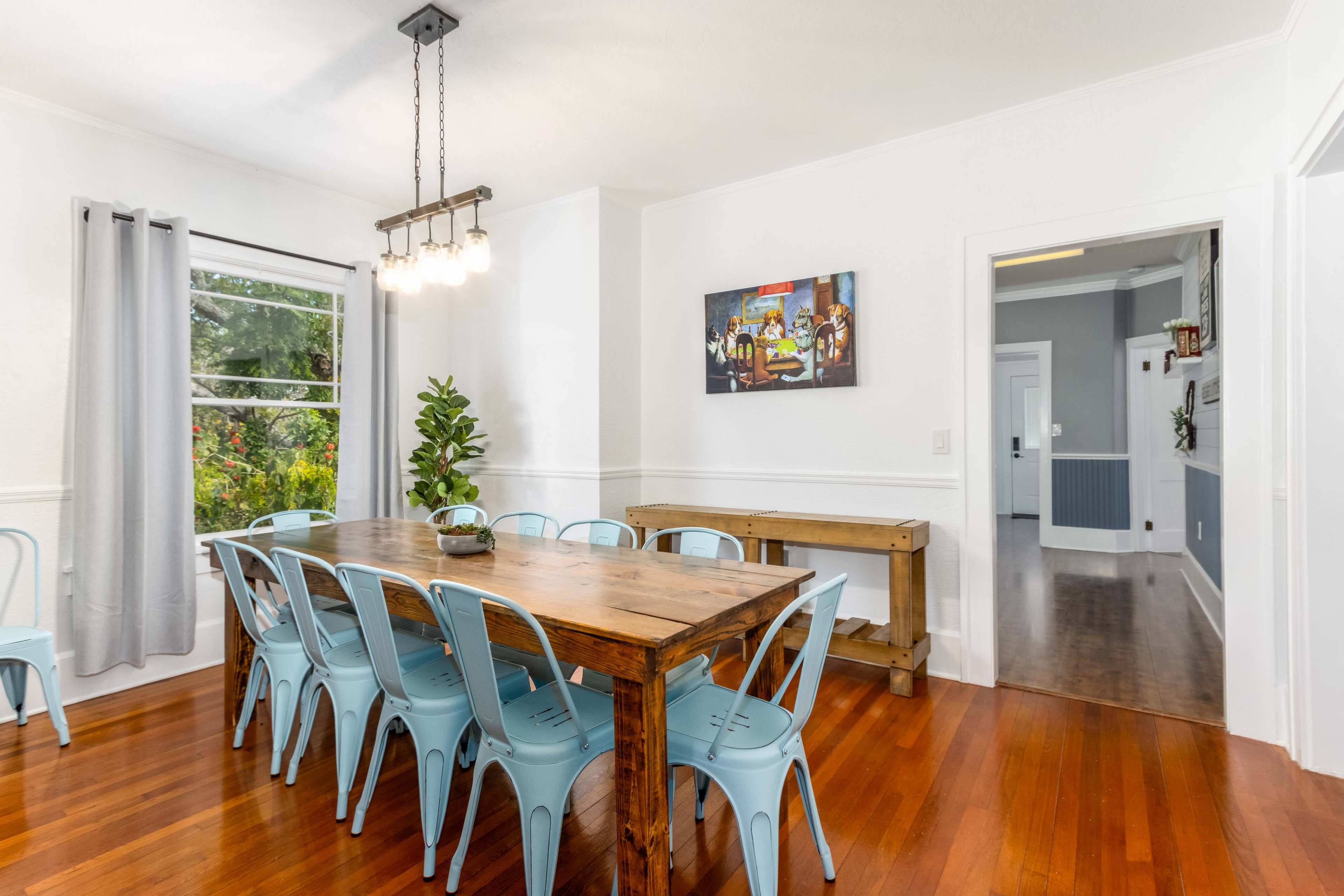 A spacious dining room features a long wooden table surrounded by blue metal chairs, with large windows allowing natural light to flood the space.