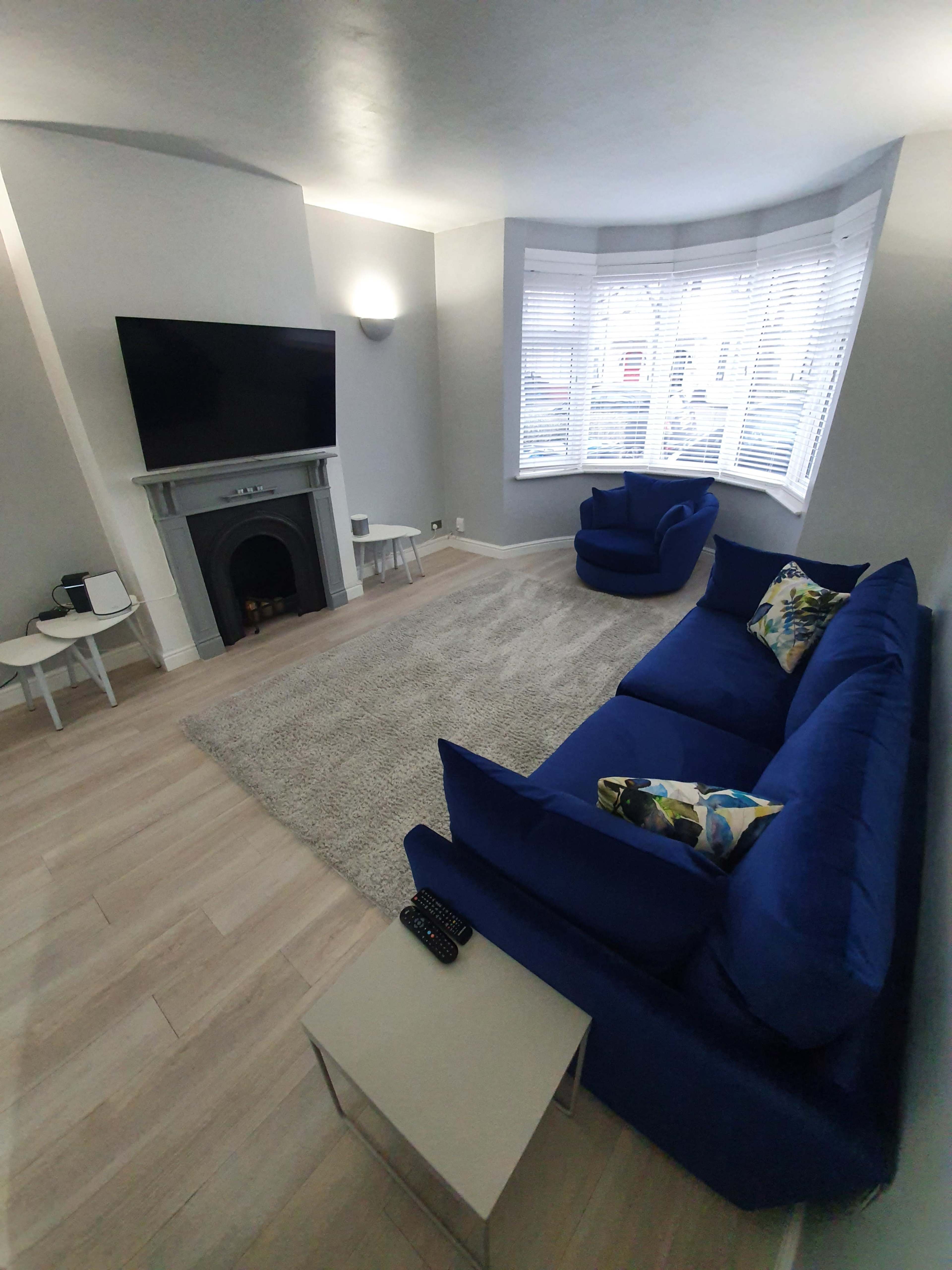 A modern living room featuring a blue sofa, two blue armchairs, a fireplace, and a large television mounted on a wall with ample natural light coming through bay windows.