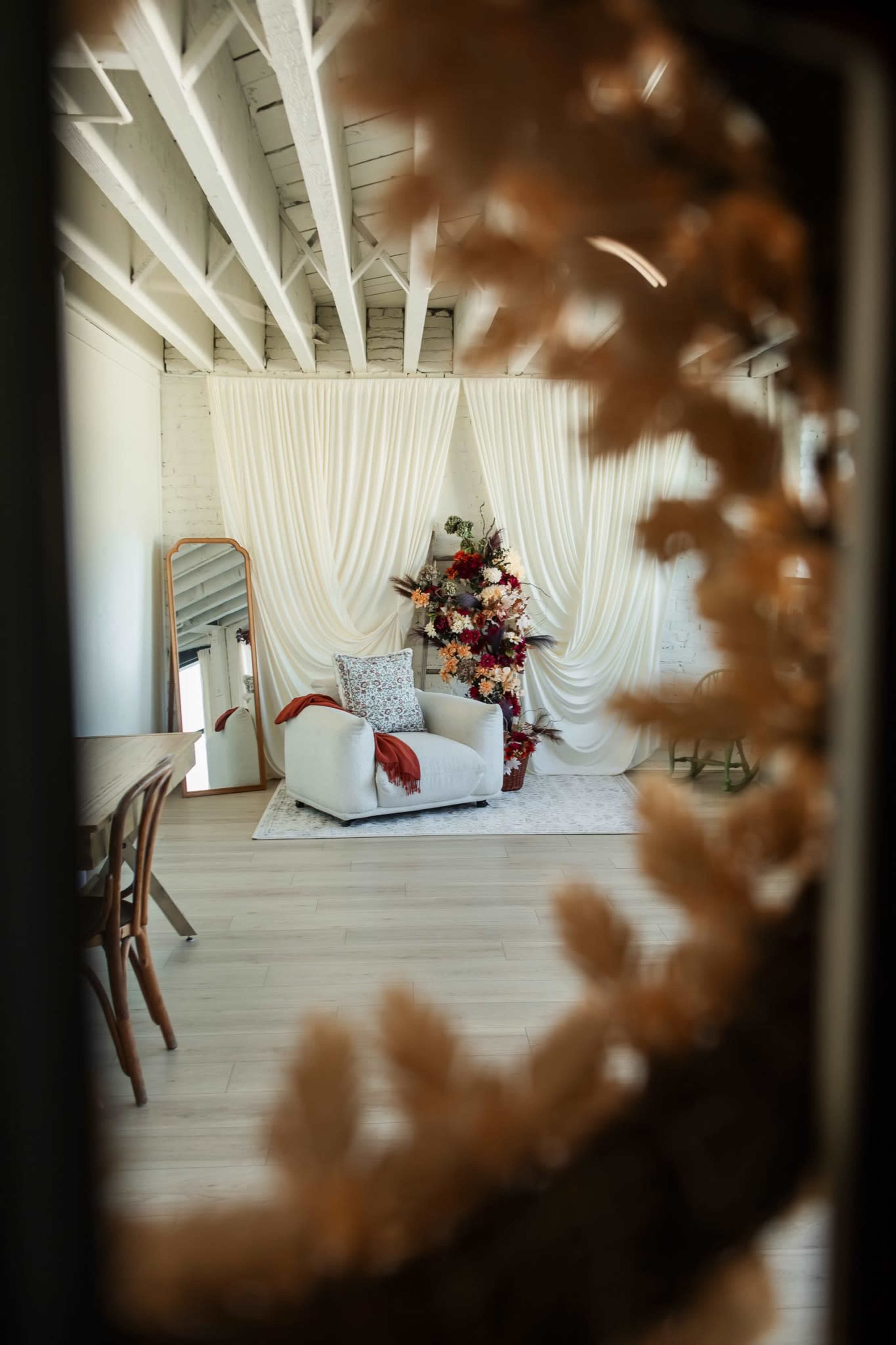 The image shows a bright, modern room featuring a white couch with a red throw blanket, a large mirror, and a floral arrangement against a backdrop of draped fabric.