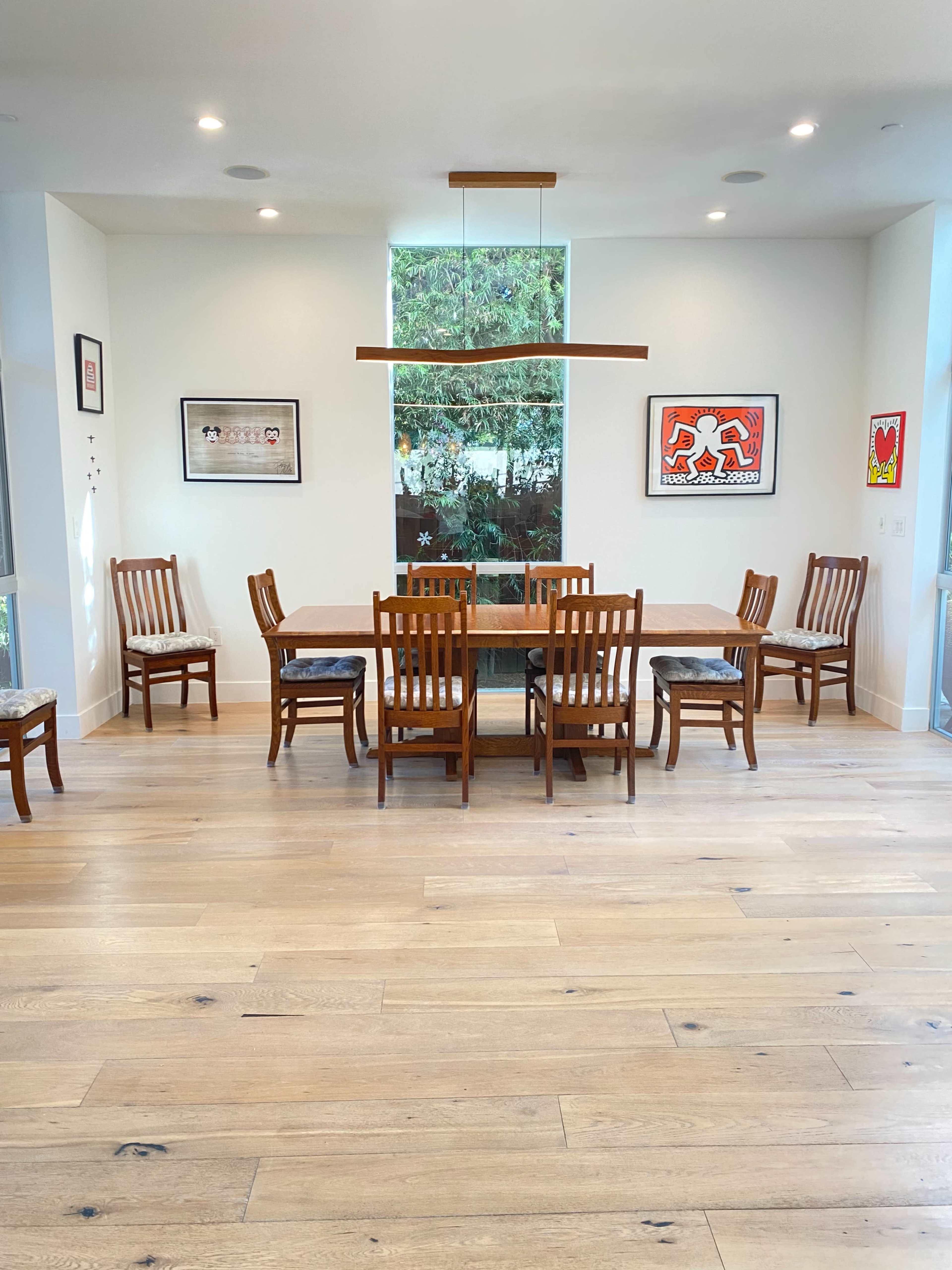 A dining area featuring a wooden table surrounded by six chairs, with art on the walls and large windows allowing natural light.