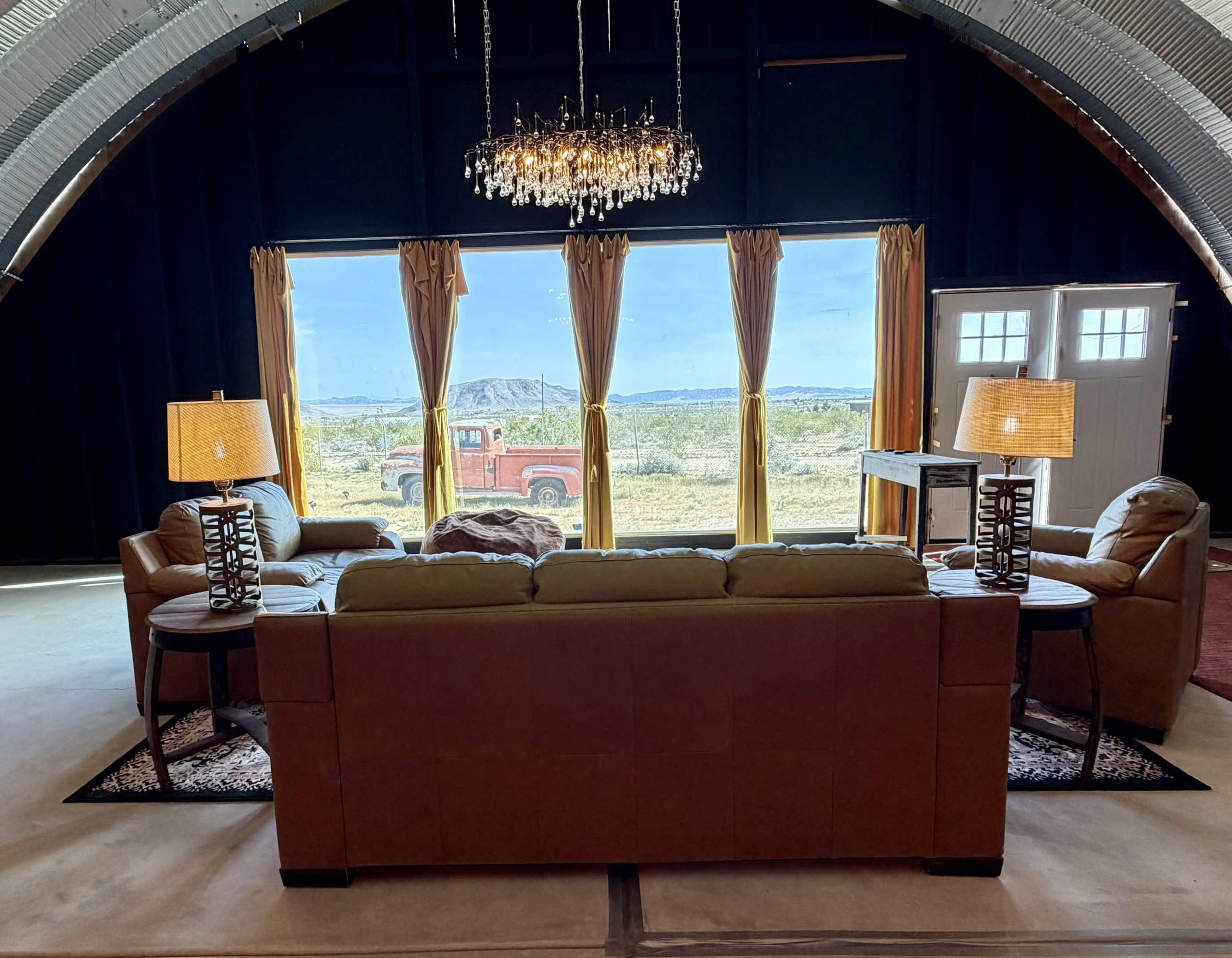 The image shows a modern living room with a brown couch, two armchairs, and large windows overlooking a desert landscape with a red truck parked outside.