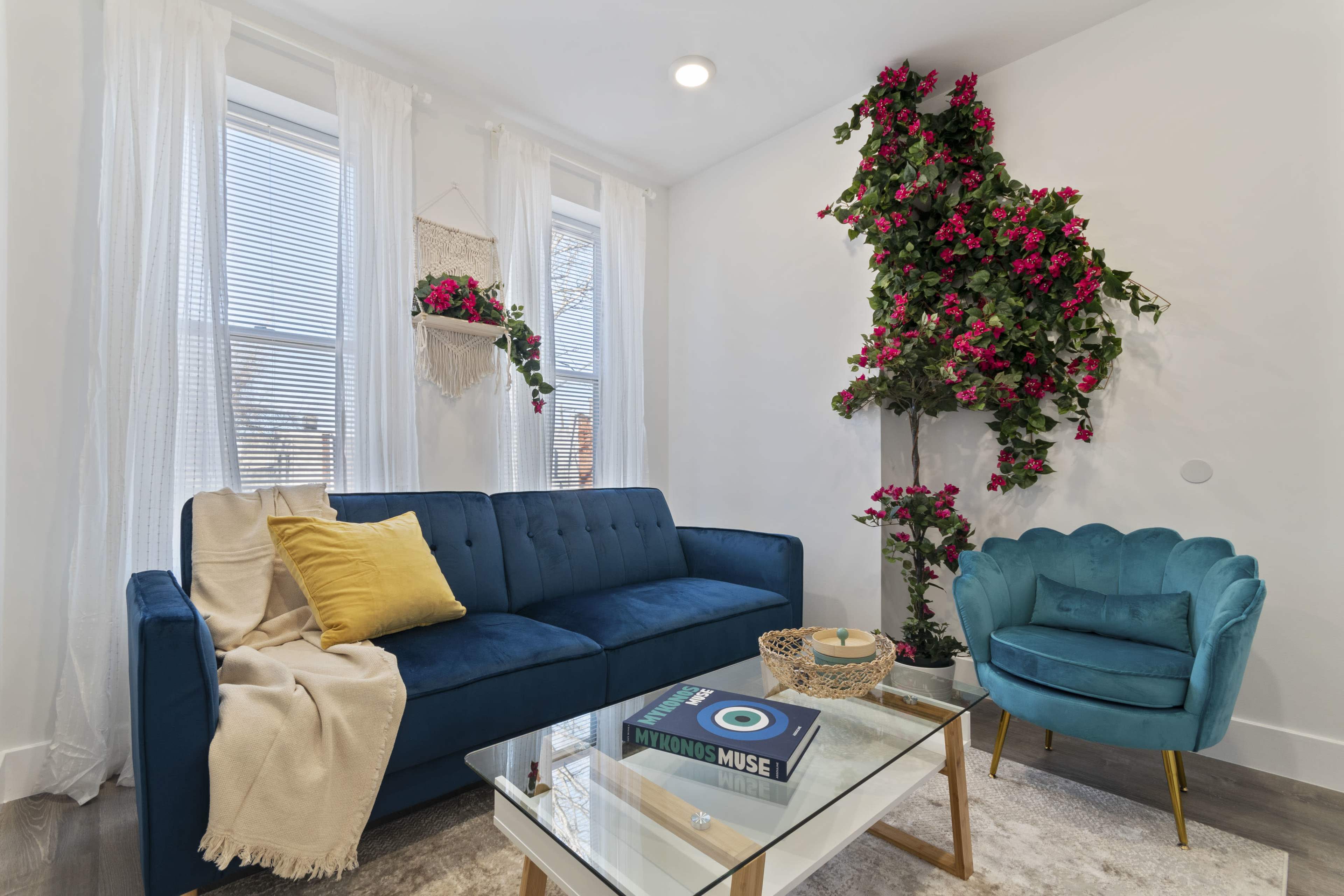 A cozy living room with a blue sofa, a light blue armchair, a glass coffee table, and decorative plants on the wall.