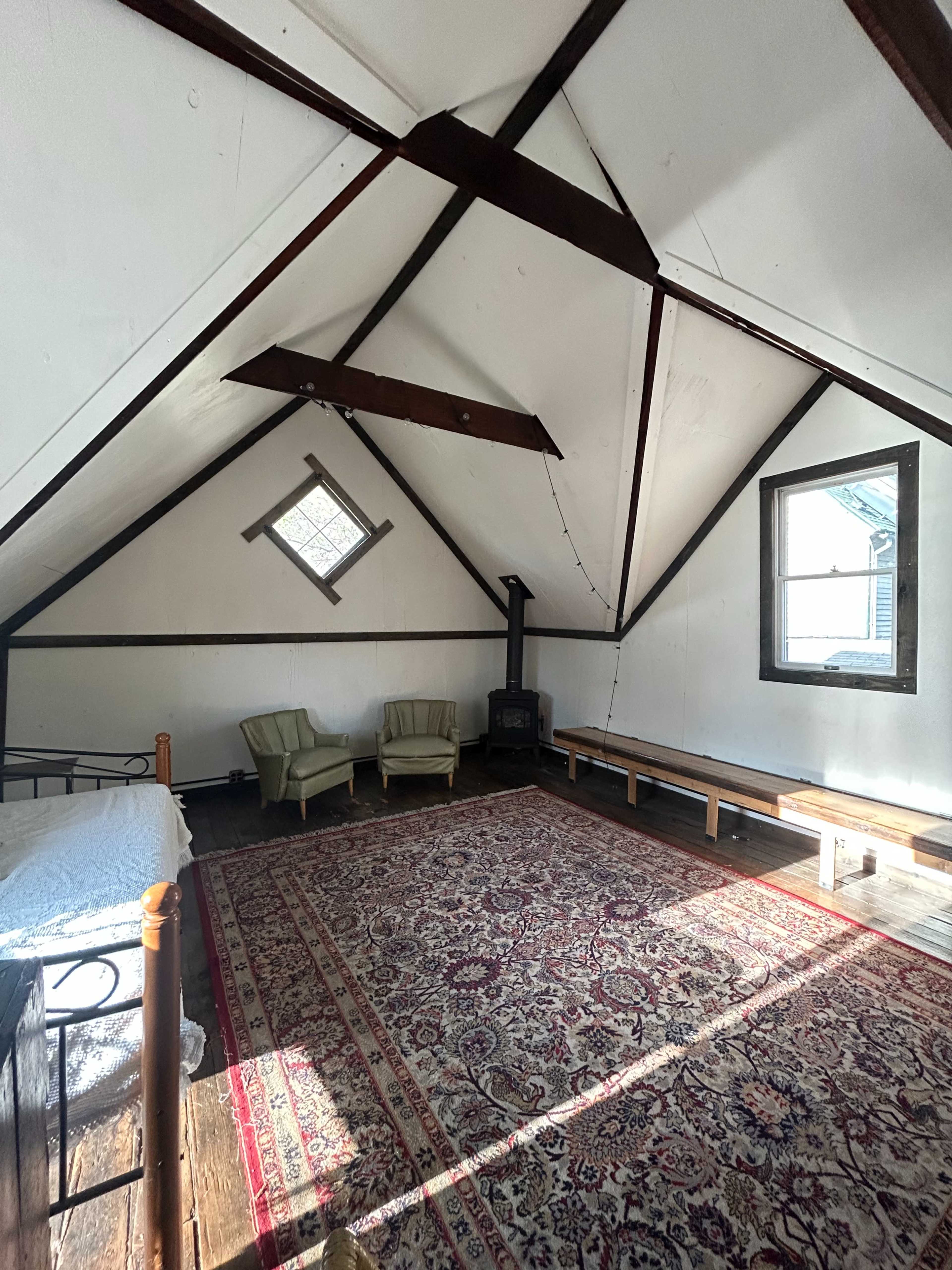 The image shows a spacious attic room with a sloped ceiling, two green chairs, a wood stove, and a large area rug on the wooden floor.
