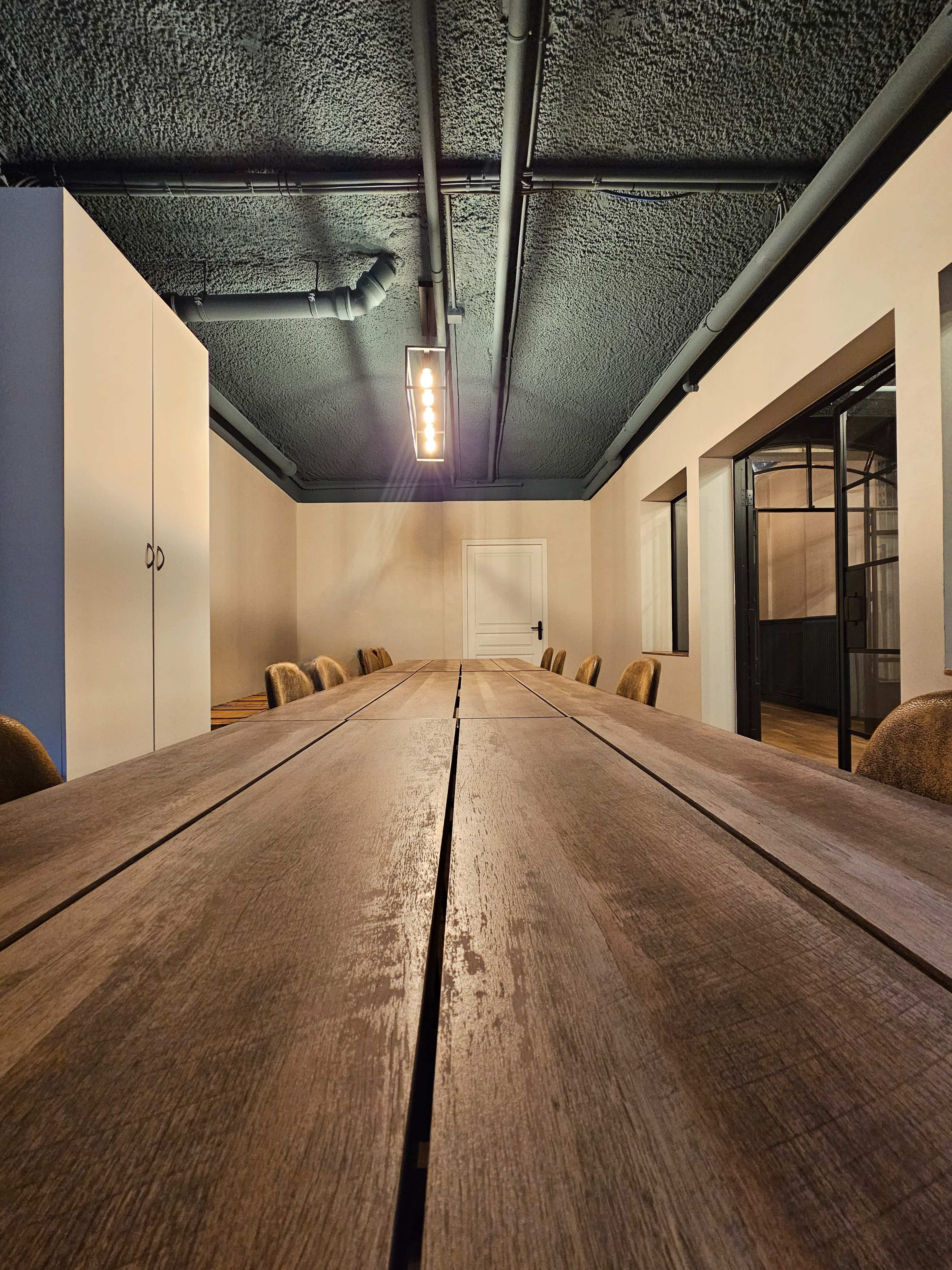 A spacious conference room featuring a long wooden table surrounded by chairs and a minimalist design with dark ceiling beams.