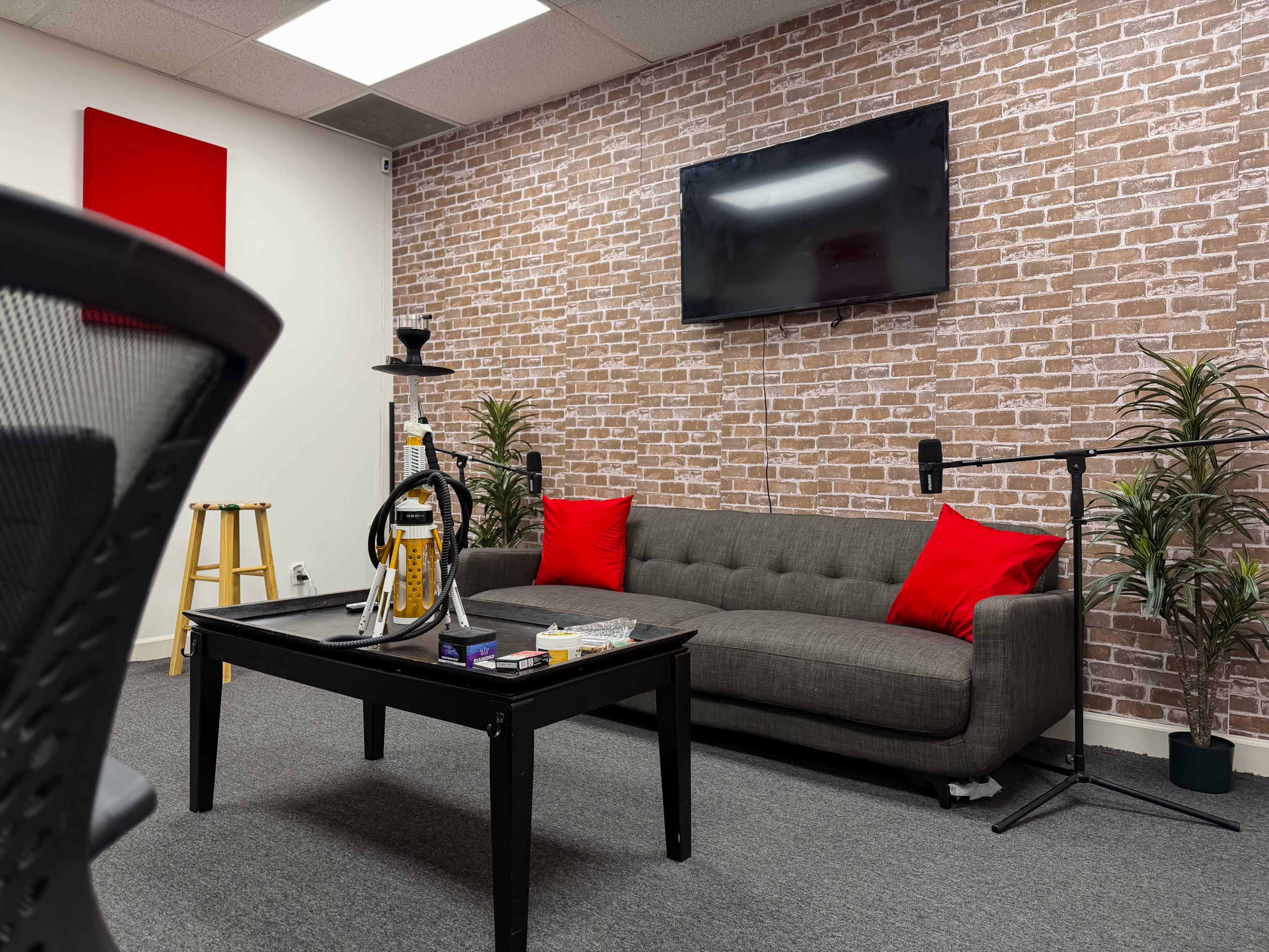 The image shows a modern lounge area with a gray sofa adorned with red cushions, a black table, and a wall featuring a brick pattern and a mounted TV.