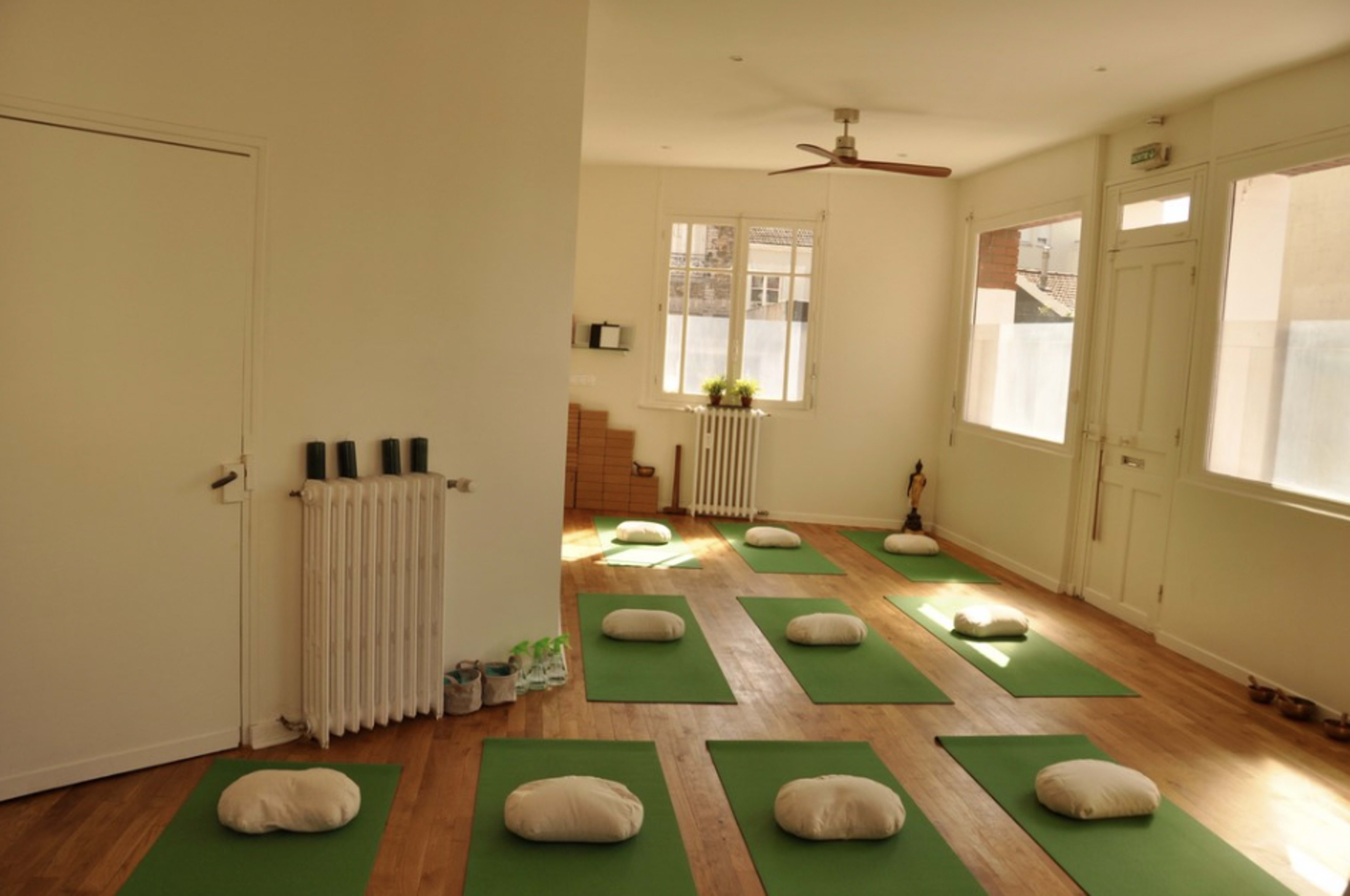 A yoga studio with green mats arranged on a wooden floor, each mat accompanied by a white cushion, and natural light entering through large windows.