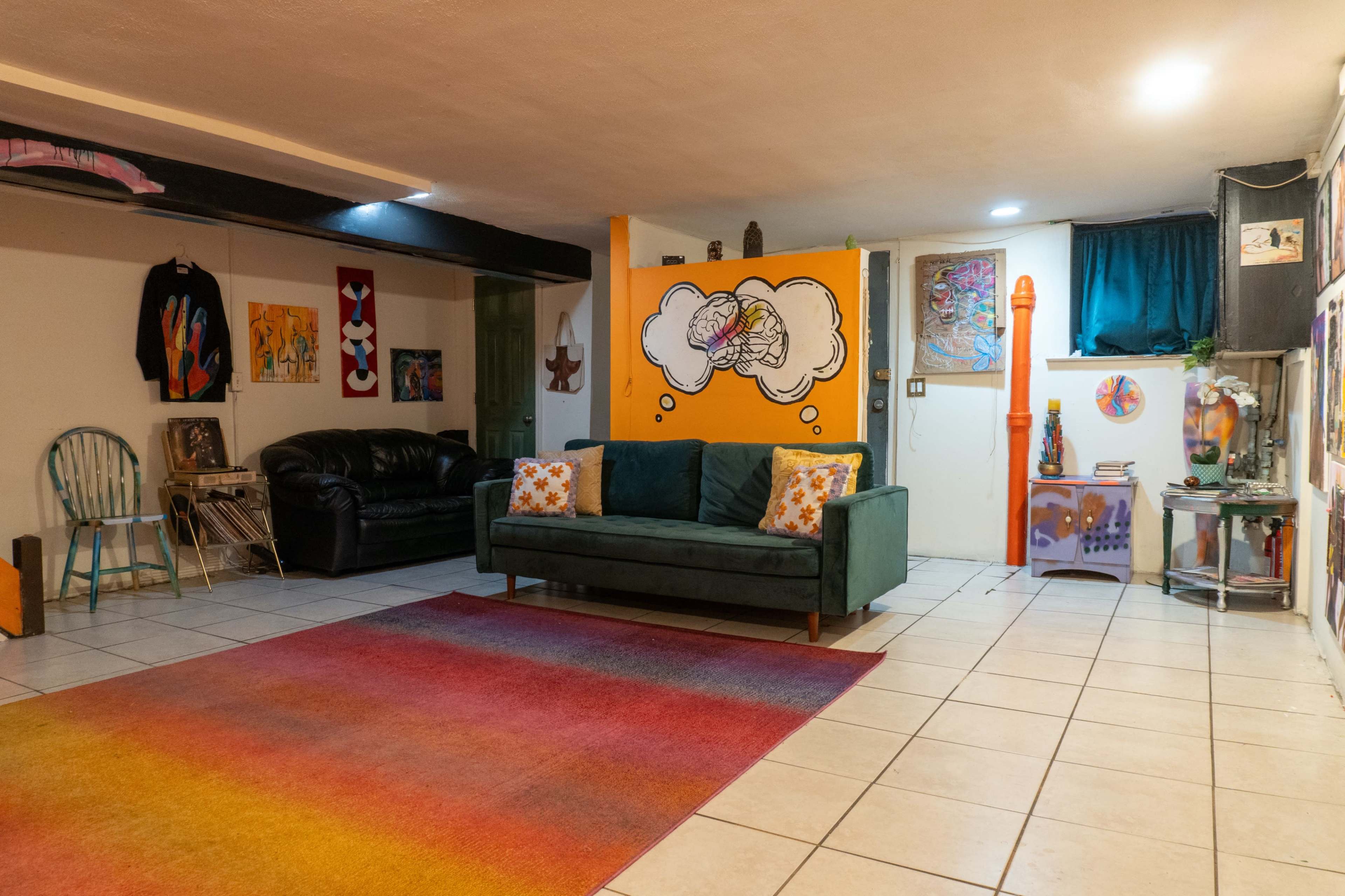 The image shows a colorful basement living area featuring a green couch with patterned cushions, a large area rug, various wall artworks, and a bookshelf.