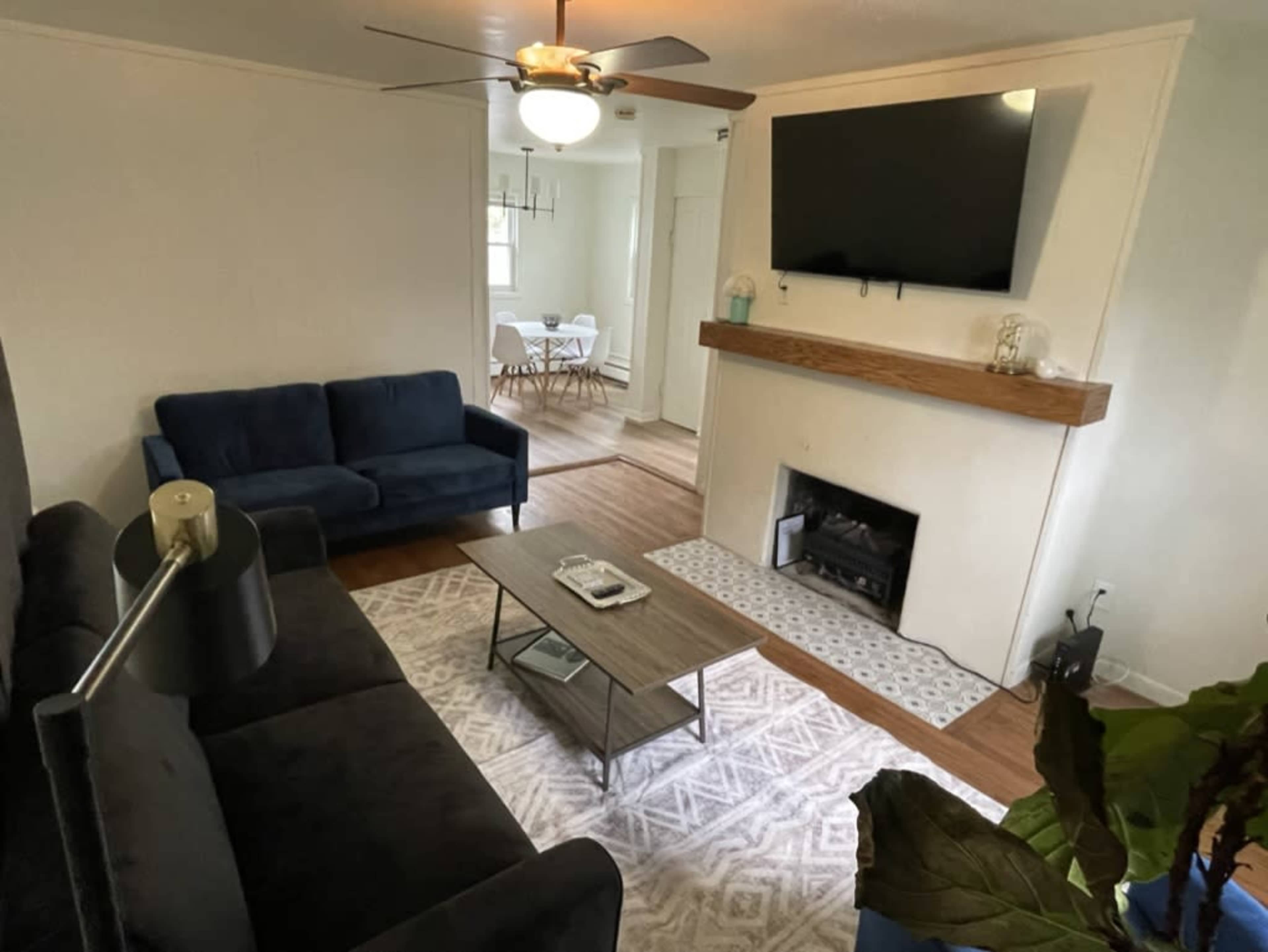 The image shows a living room featuring a dark blue sofa, a coffee table, a wall-mounted television, and a small fireplace with a wooden mantel.