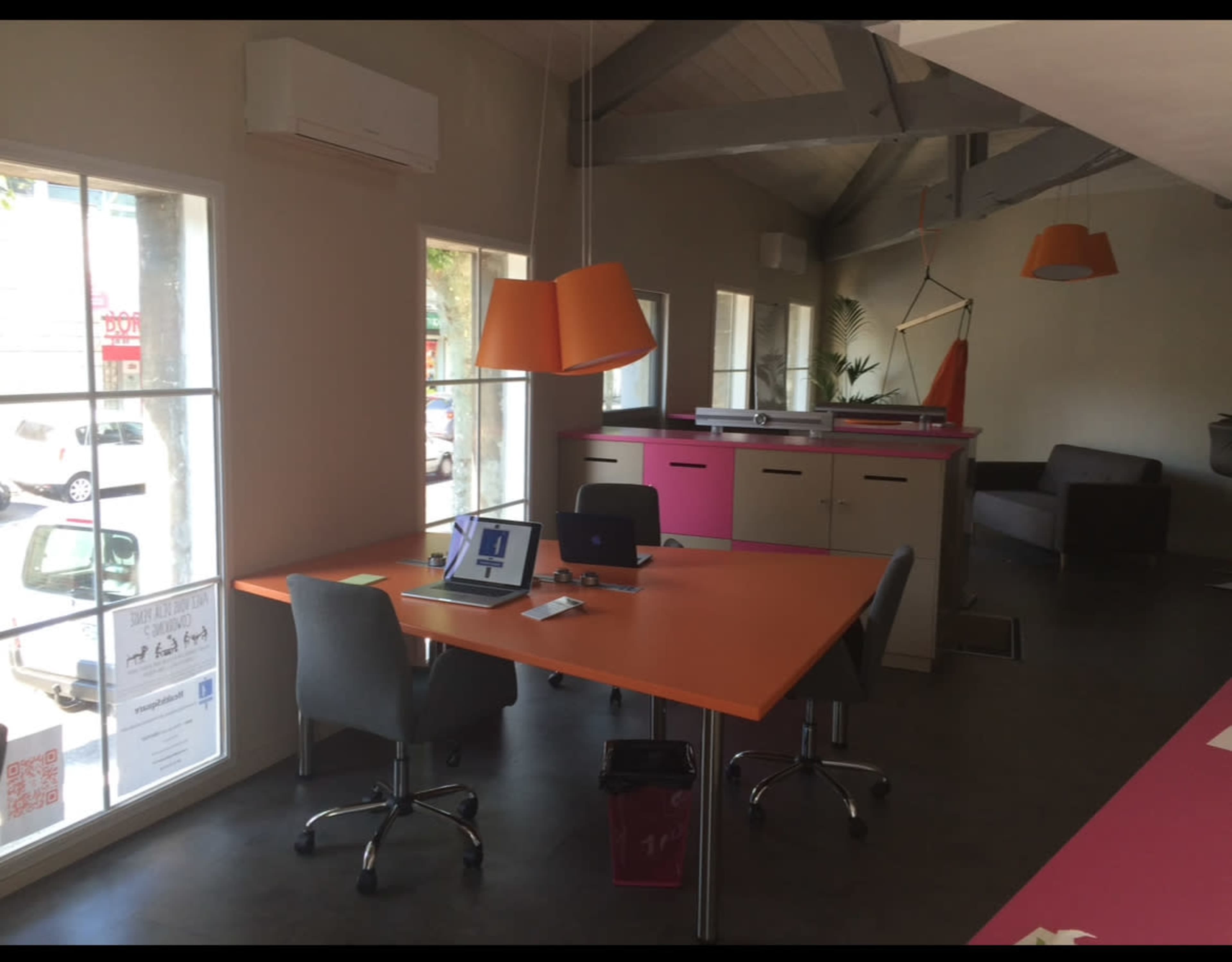 The image shows a modern office space with large windows, a bright orange table, gray chairs, and colorful accents, including an orange pendant light and pink design elements.