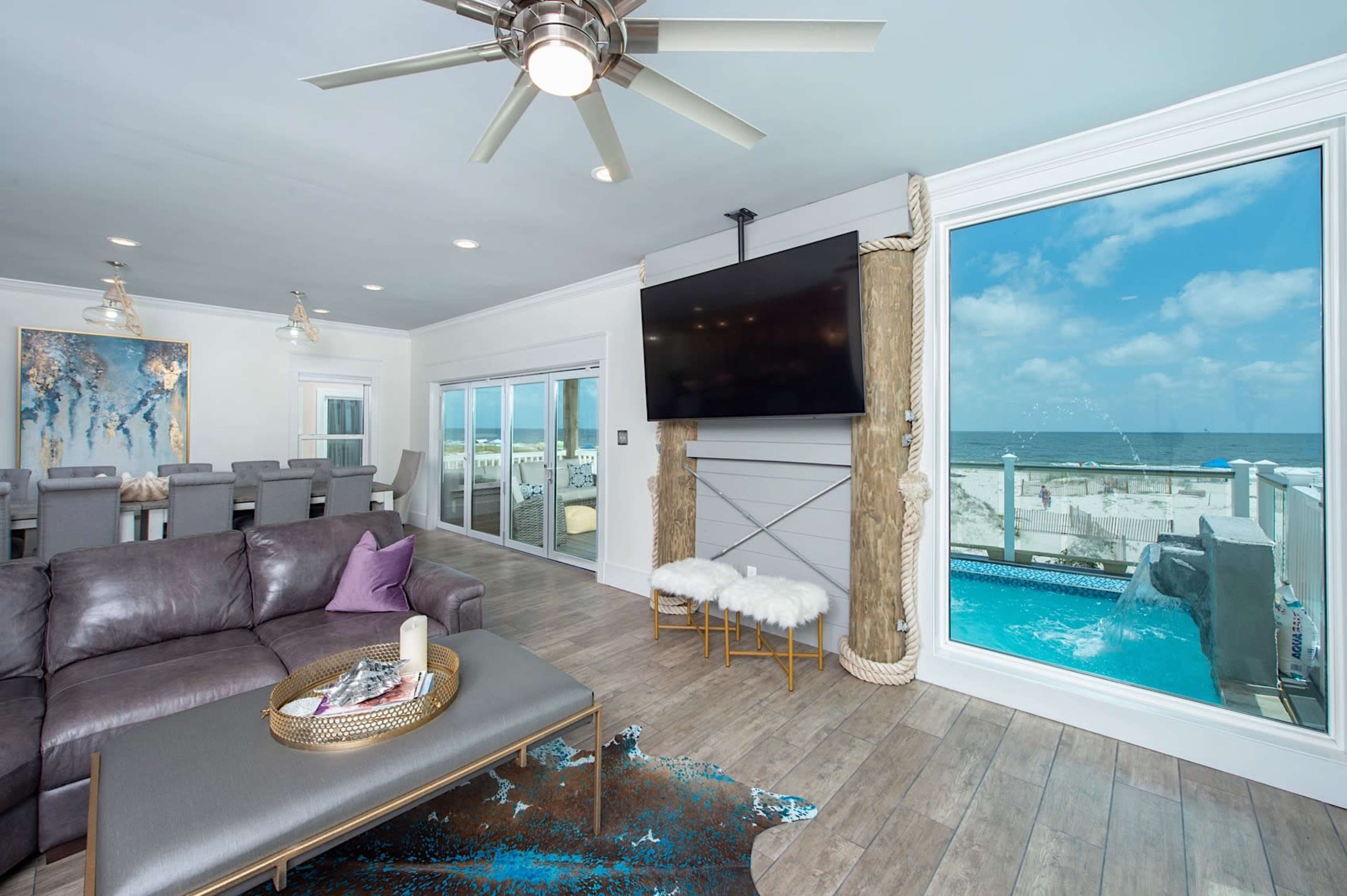 A modern living room features a large window overlooking a beach view, a dark leather sofa, and a wall-mounted television.