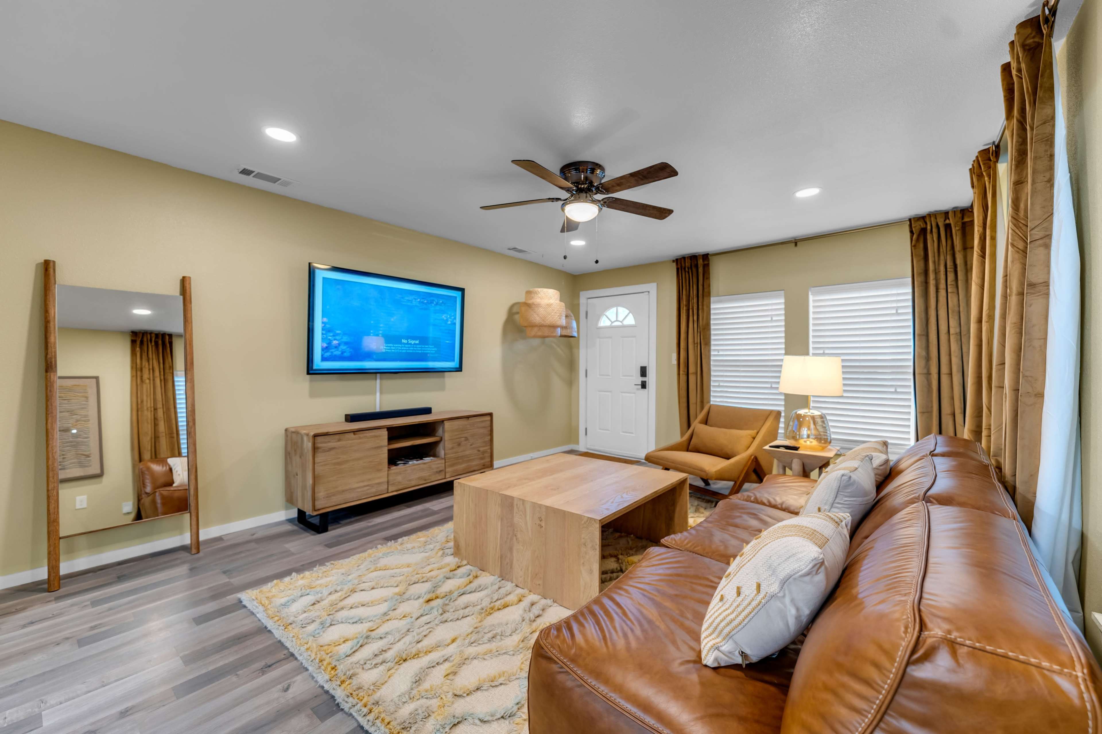 A modern living room features a leather sofa, wooden coffee table, and a television mounted on the wall, with a front door and window letting in natural light.