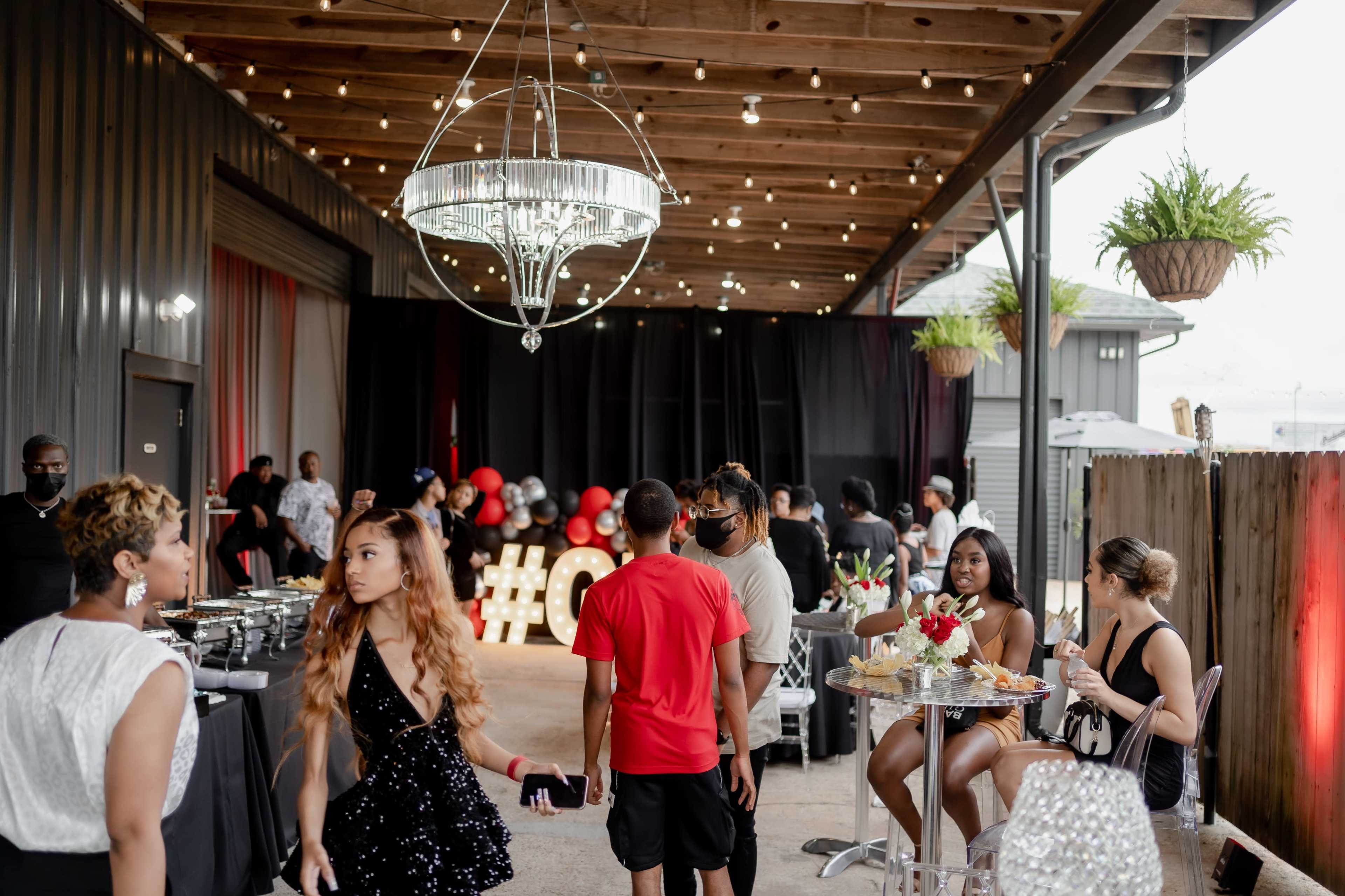 A lively gathering takes place in a decorated indoor space featuring tables, a chandelier, and a backdrop of balloons.