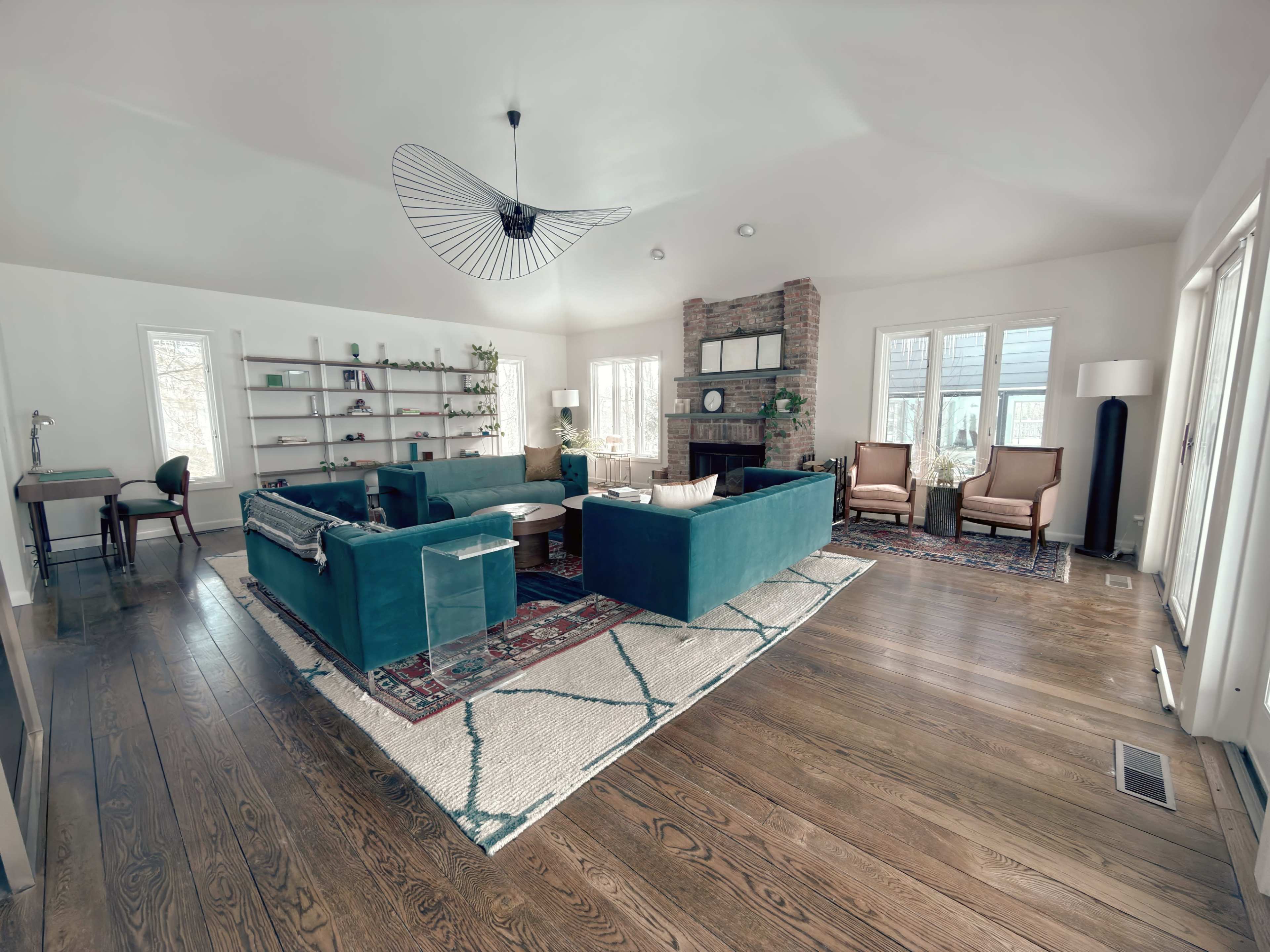 The image shows a modern living room featuring a teal sectional sofa, a stone fireplace, bookshelves, and wooden flooring, with large windows allowing natural light to enter.