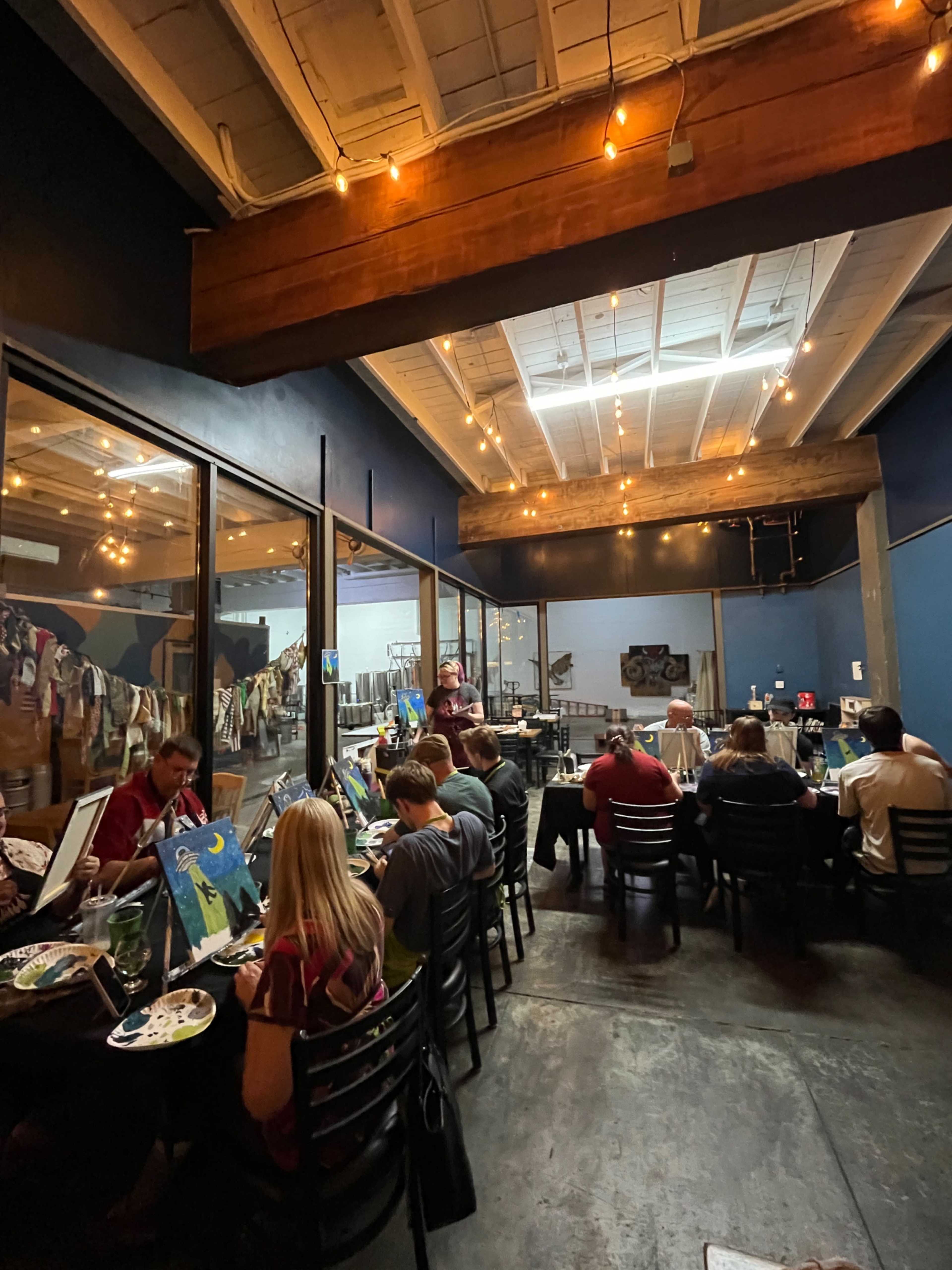 The image shows a restaurant interior where diners are eating and painting, with colorful artwork displayed along the walls under warm lighting.